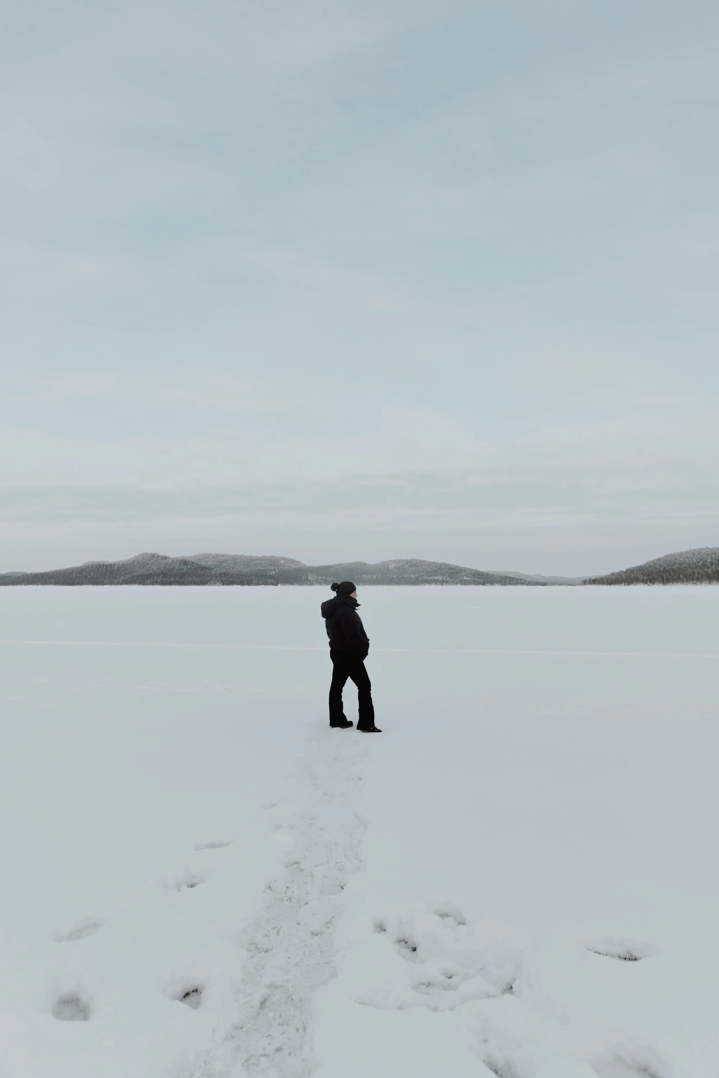 Une personne en vêtements d'hiver debout sur un lac gelé, avec des montagnes en arrière-plan, dans un paysage enneigé et froid.