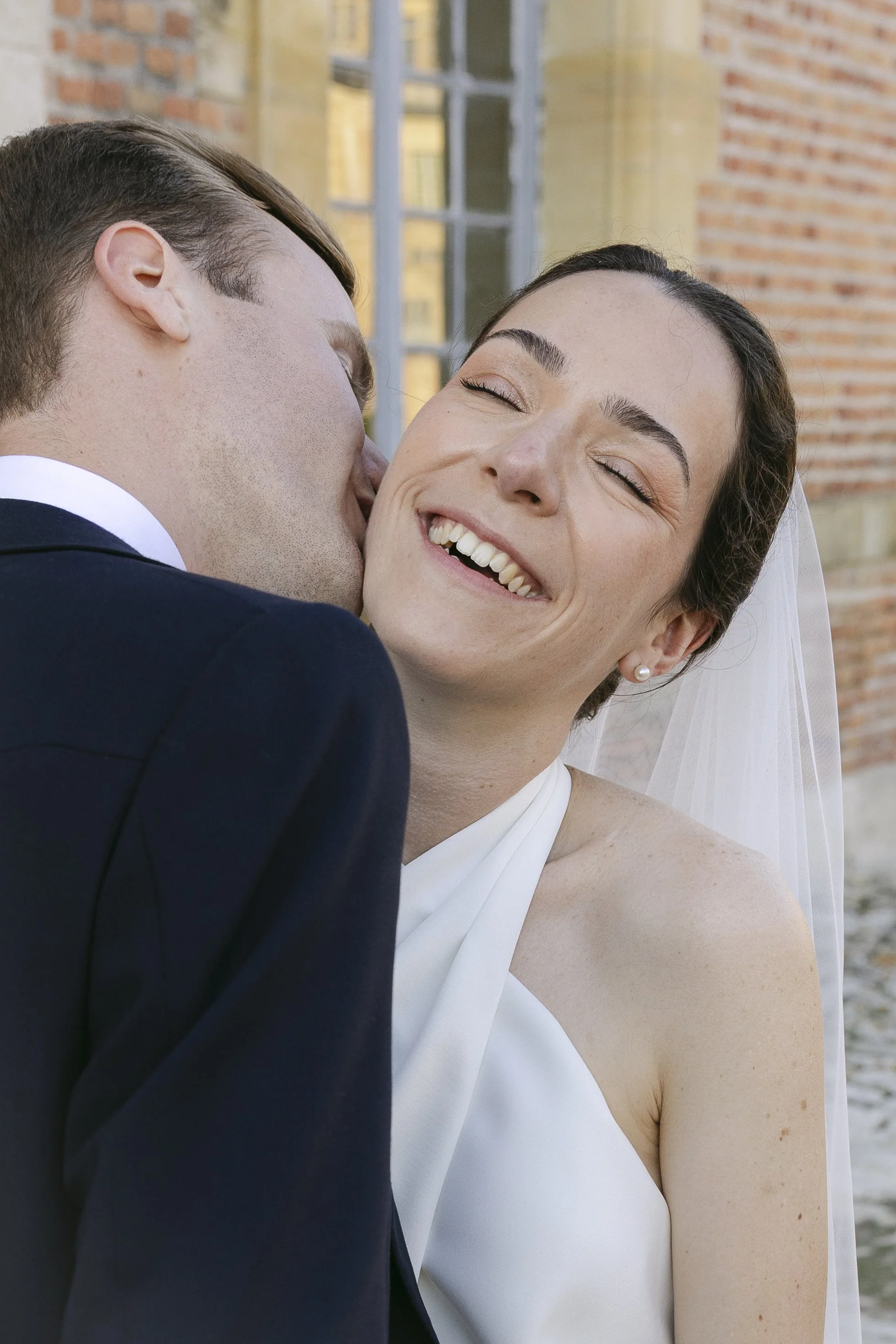 Un couple de mariés s'embrassant avec une expression de bonheur, en extérieur, près d'une bâtisse en briques.