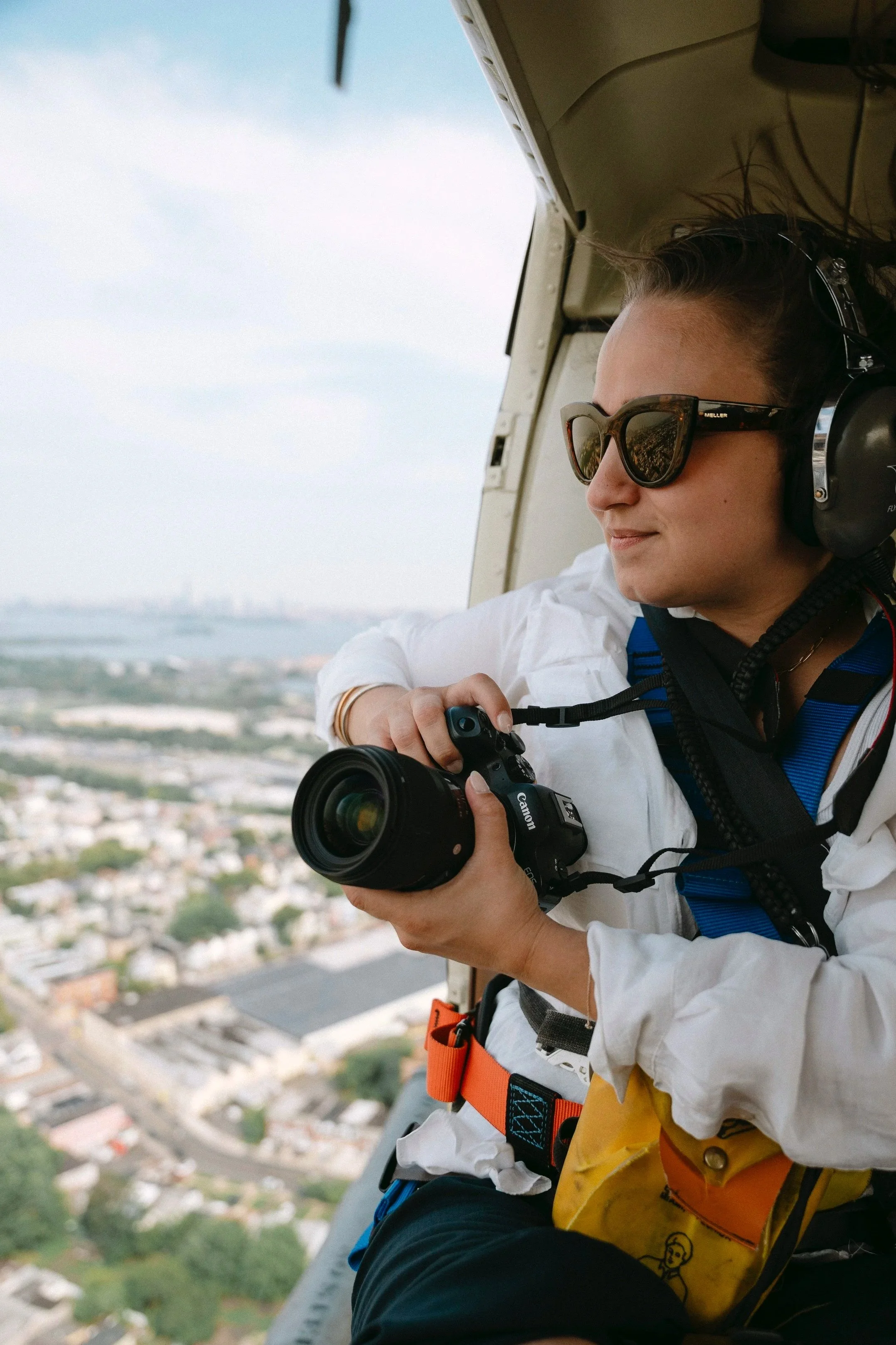 Une femme portant des lunettes de soleil et un casque d'écoute, tenant un appareil photo, regarde à l'extérieur d'un hélicoptère lors d'un vol au-dessus d'une ville.
