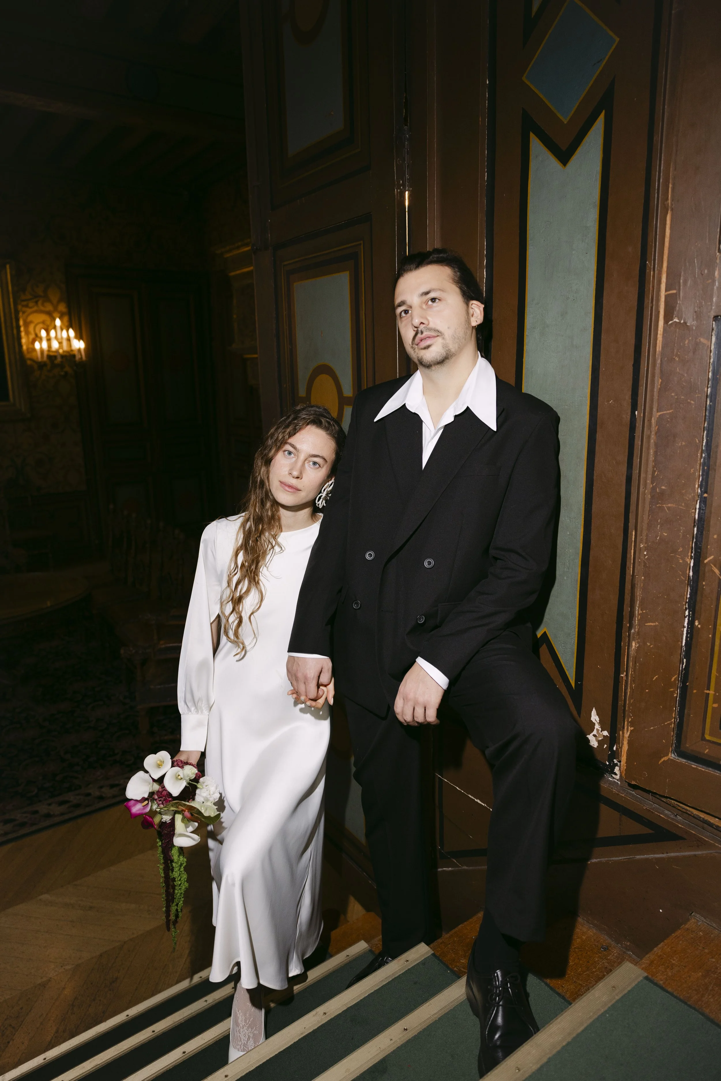 Un couple posant dans un intérieur élégant. La femme porte une robe blanche et tient un bouquet de fleurs, tandis que l'homme porte un costume noir avec une chemise blanche.