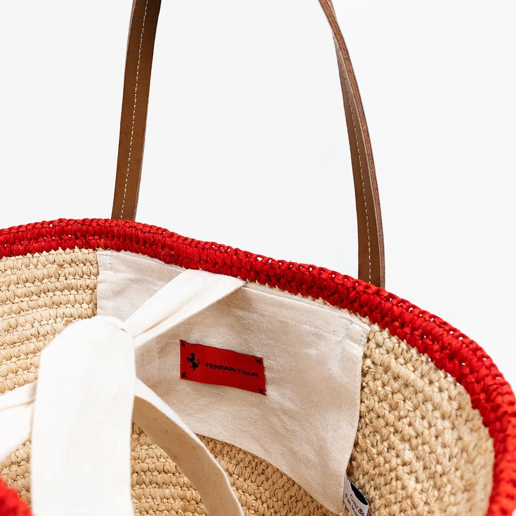 Une sac en osier avec des poignées en cuir marron, bordure rouge en crochet, intérieur en tissu blanc avec un label rouge "FERRARI TOUR".