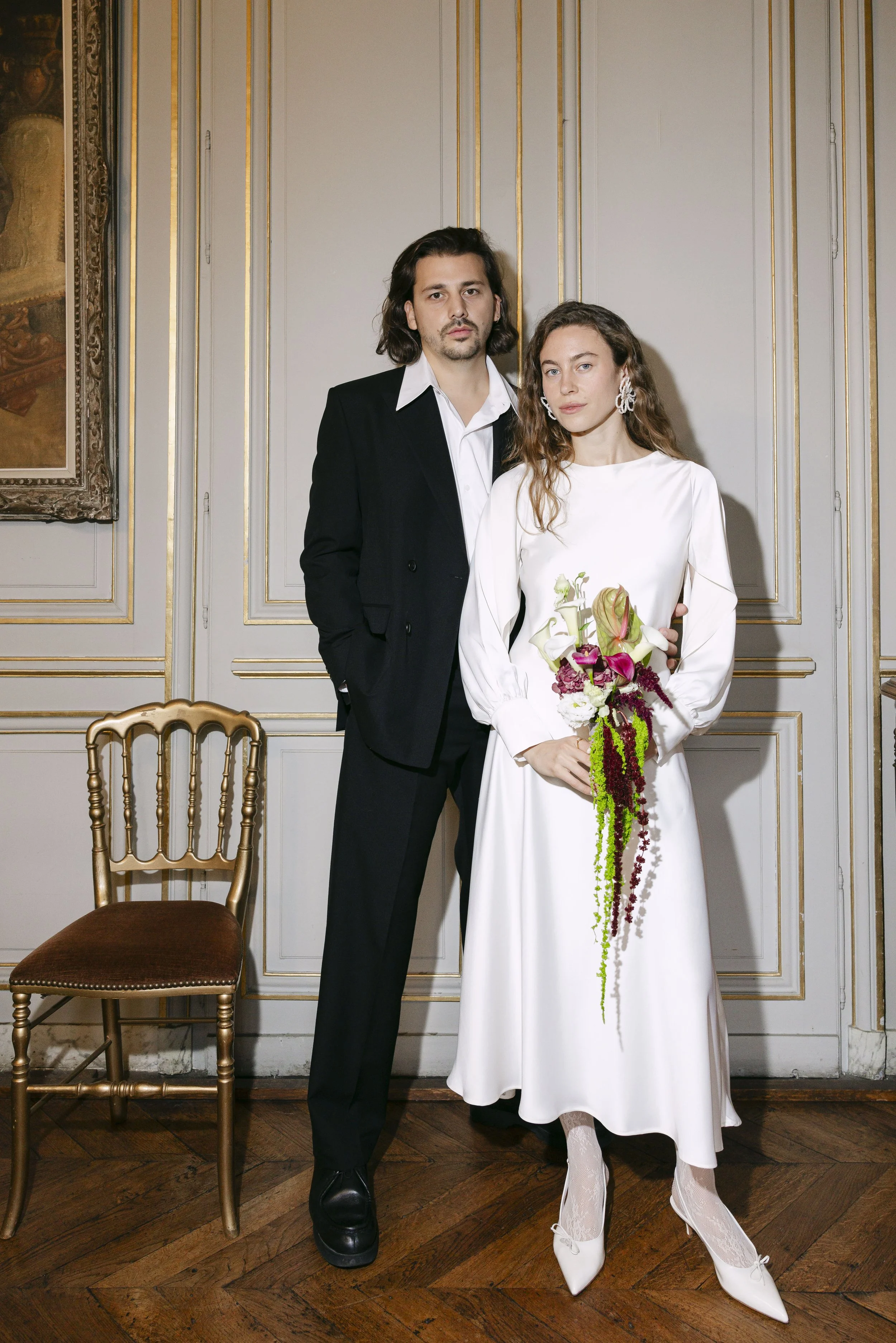 Un couple habillé pour un mariage posé dans une pièce élégante avec des murs ornés de moulures dorées, la femme porte une robe blanche, des chaussures à talons blanches et des bijoux, tenant un bouquet de fleurs colorées, l'homme porte un costume noir avec une chemise blanche.