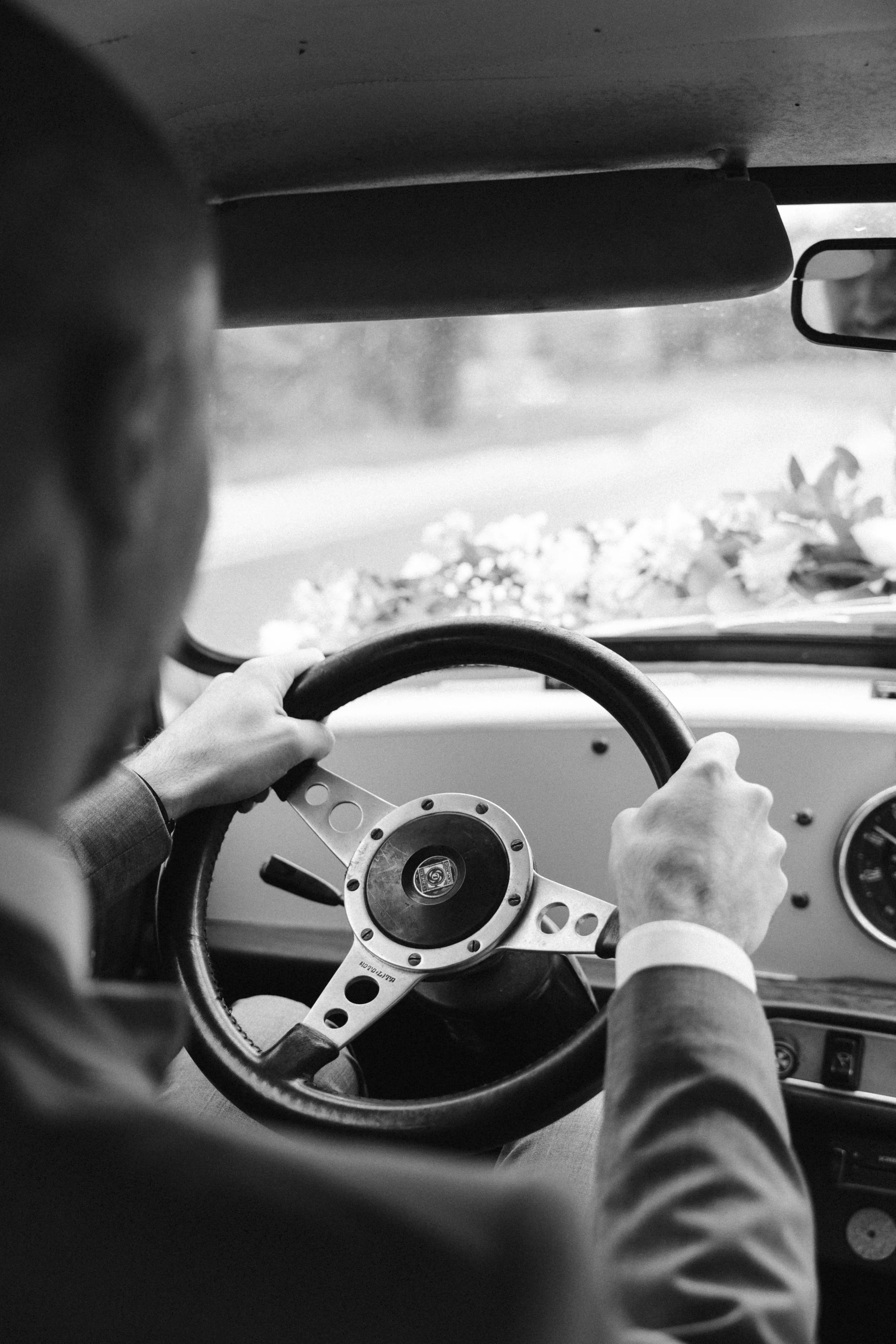 Un homme en costume conduit une voiture, vue de derrière, en noir et blanc.