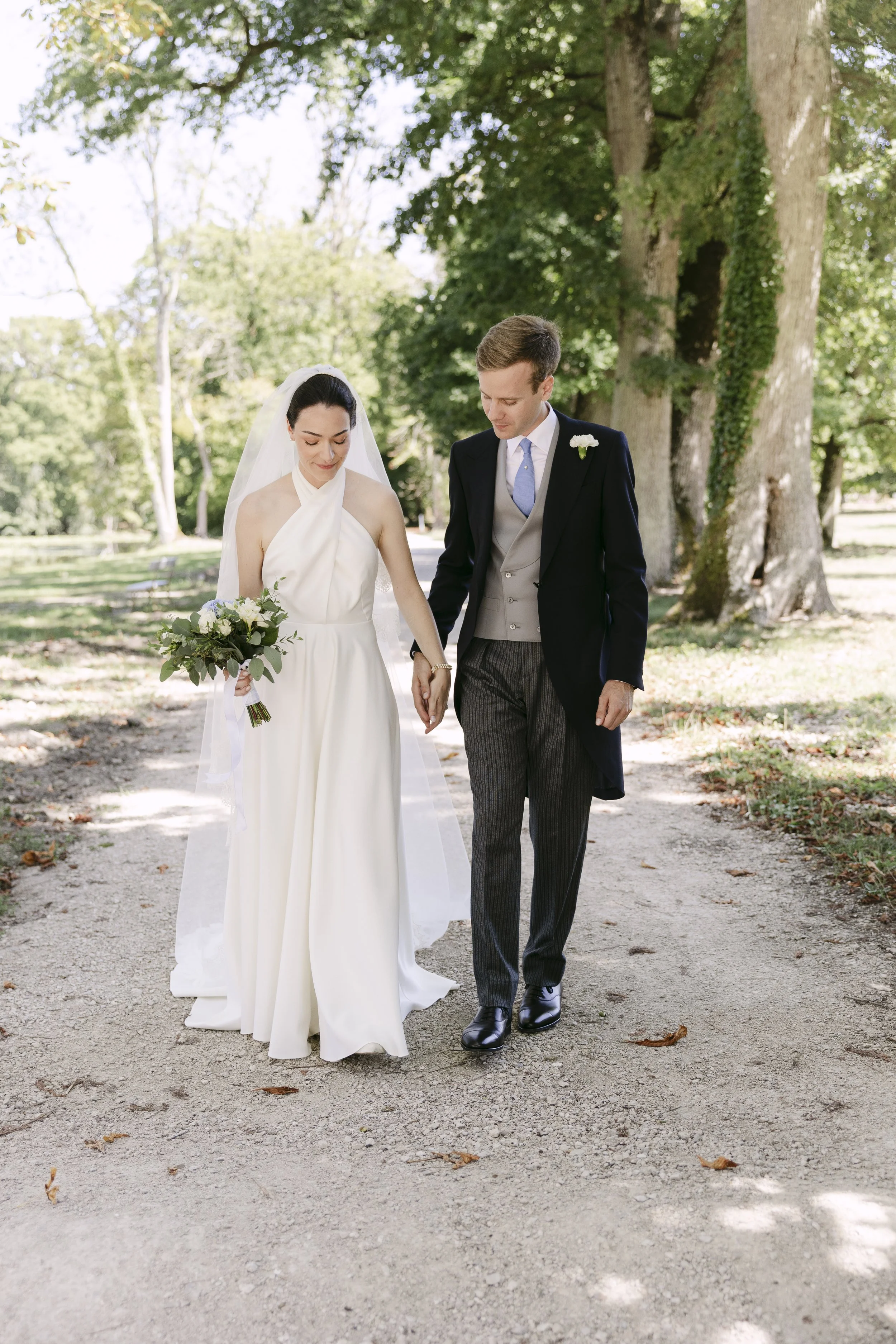 Un couple de mariés marche main dans la main dans un parc en plein air, entouré d'arbres verts, lors d'une cérémonie de mariage.