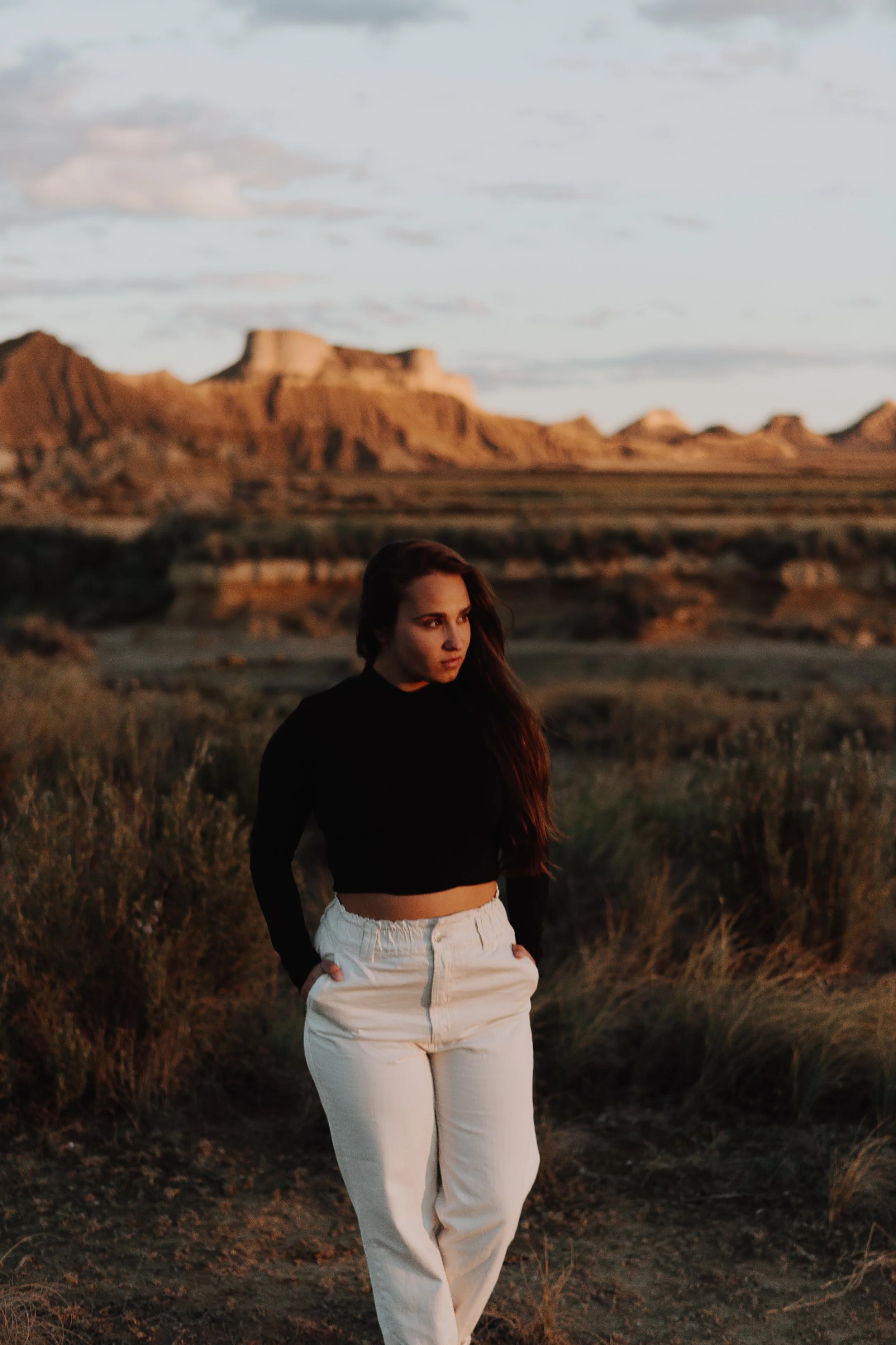 Une femme debout dans un paysage désertique avec des montagnes en arrière-plan, portant un haut noir et un pantalon blanc.