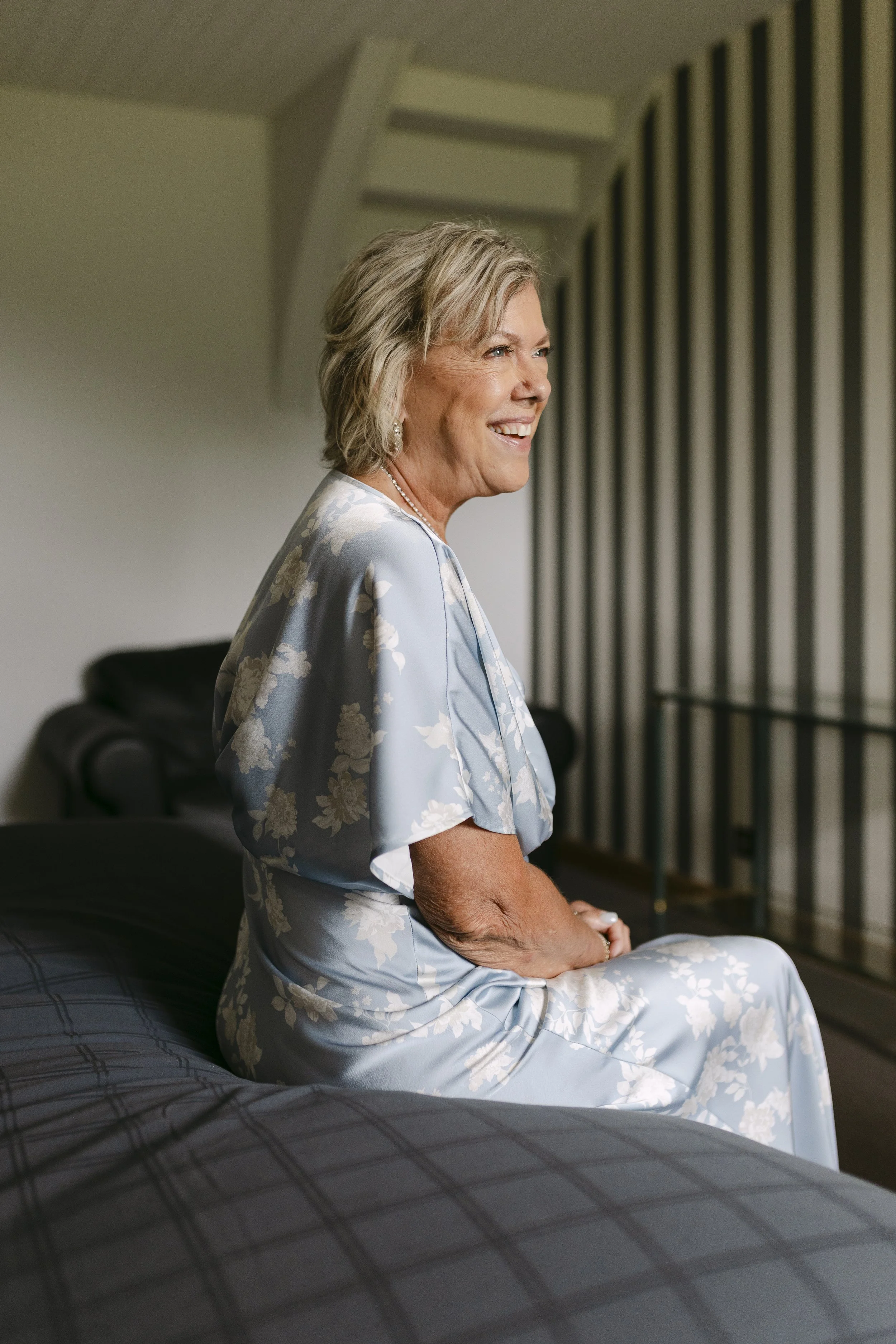 Une femme âgée souriante et habillée en robe en satin brodée de fleurs, assise sur un lit dans une pièce avec un décor minimaliste.