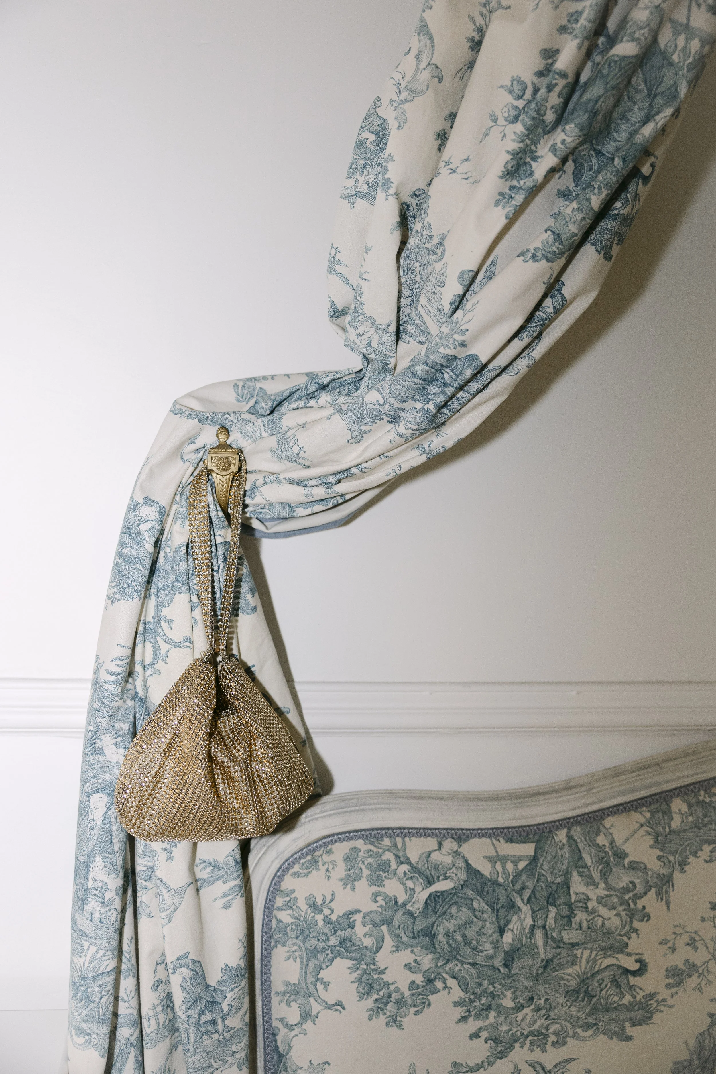 Un rideau en tissu avec un motif arlequin en teintes bleues et blanches, attaché avec une broche décorative dorée, dépassant d'une tête de lit rembourrée avec un tissu à motif toile de Jouy.