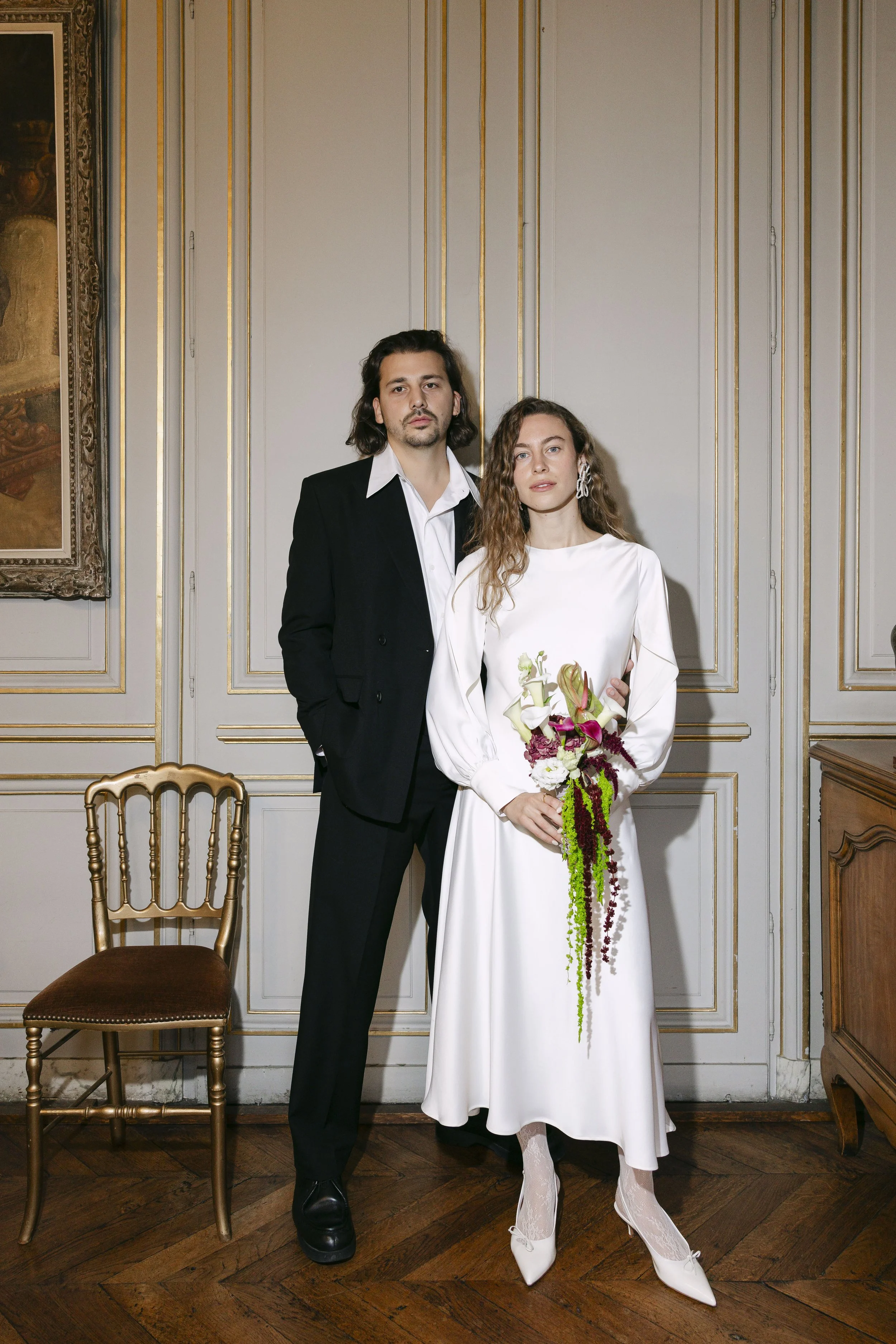 Un homme et une femme posent côte à côte dans une pièce élégante, la femme tenant un bouquet de fleurs, vêtus de vêtements formels, dans une ambiance sophistiquée.
