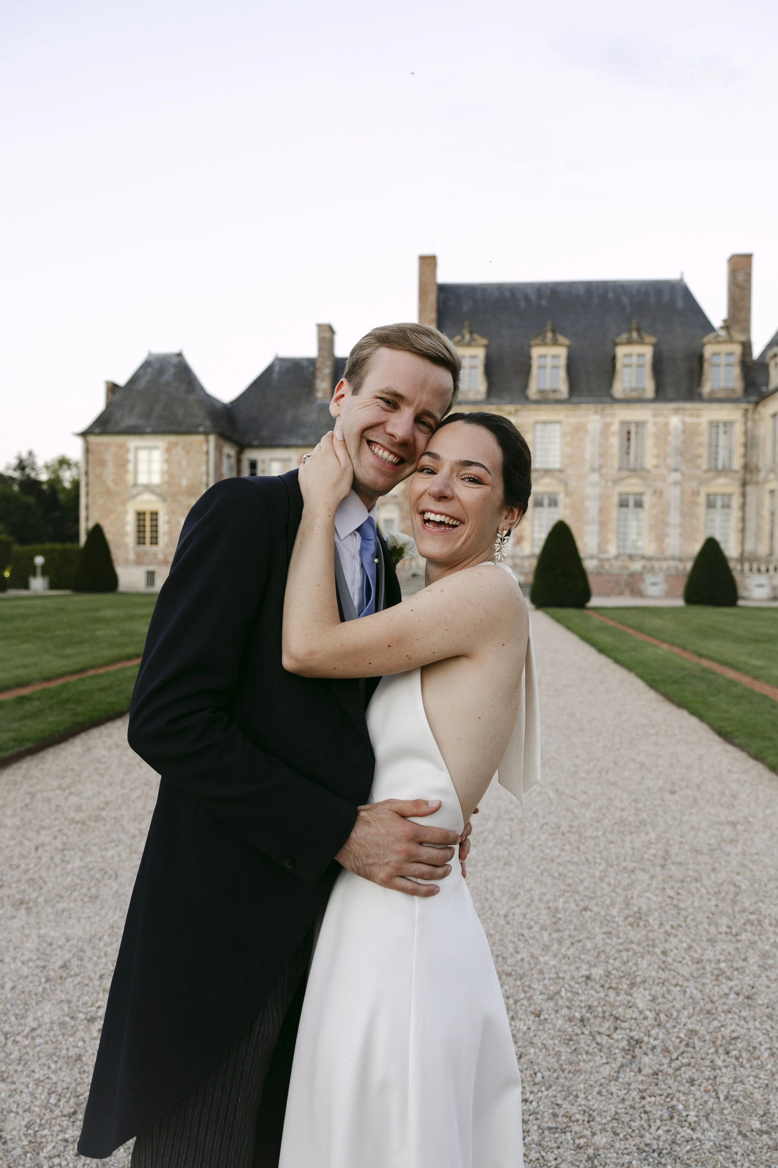 Un couple de mariés souriants en tenue de mariage, embrassant devant un grand château en pierre.