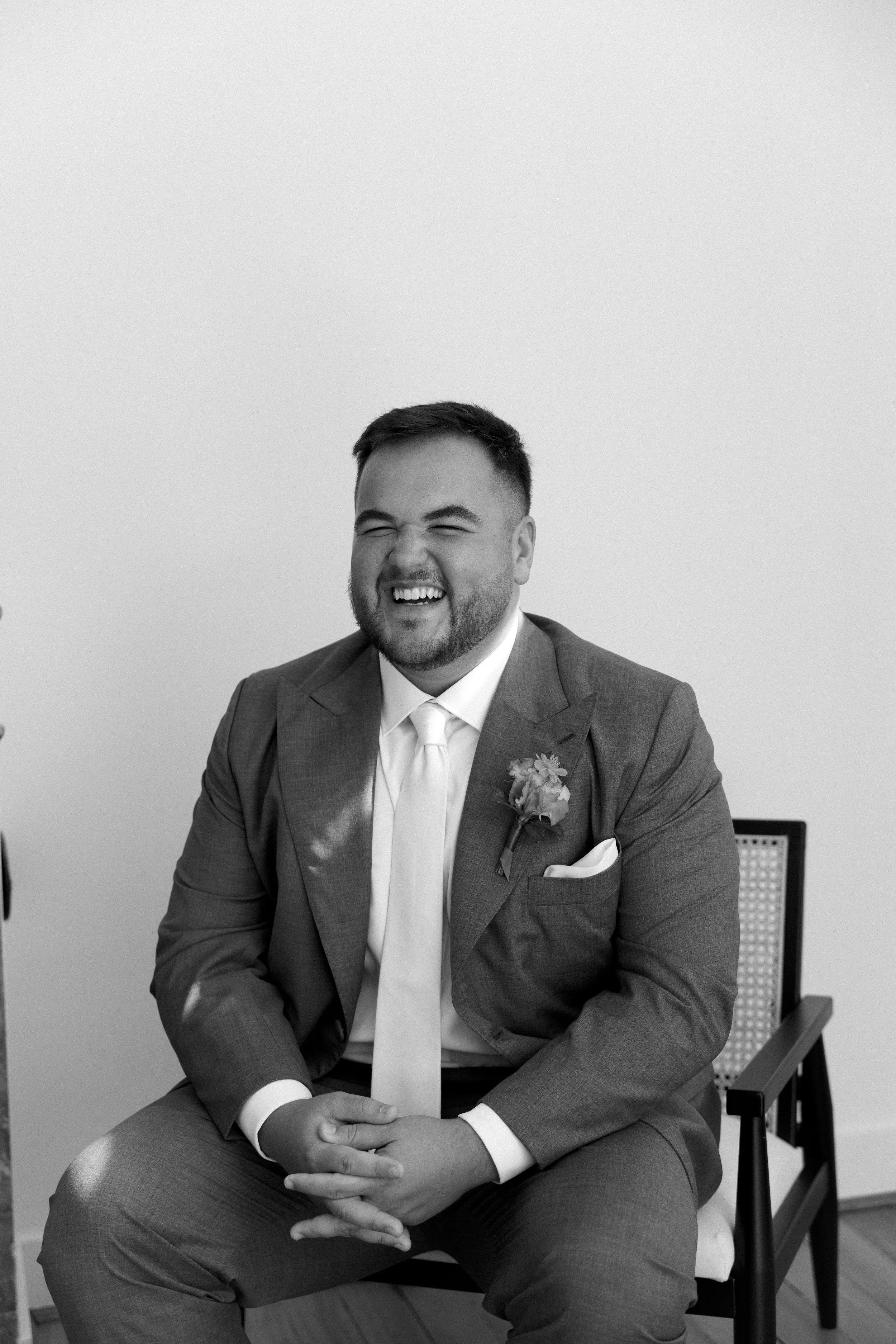 Homme souriant en costume de mariage avec une boutonnière, assis sur une chaise.