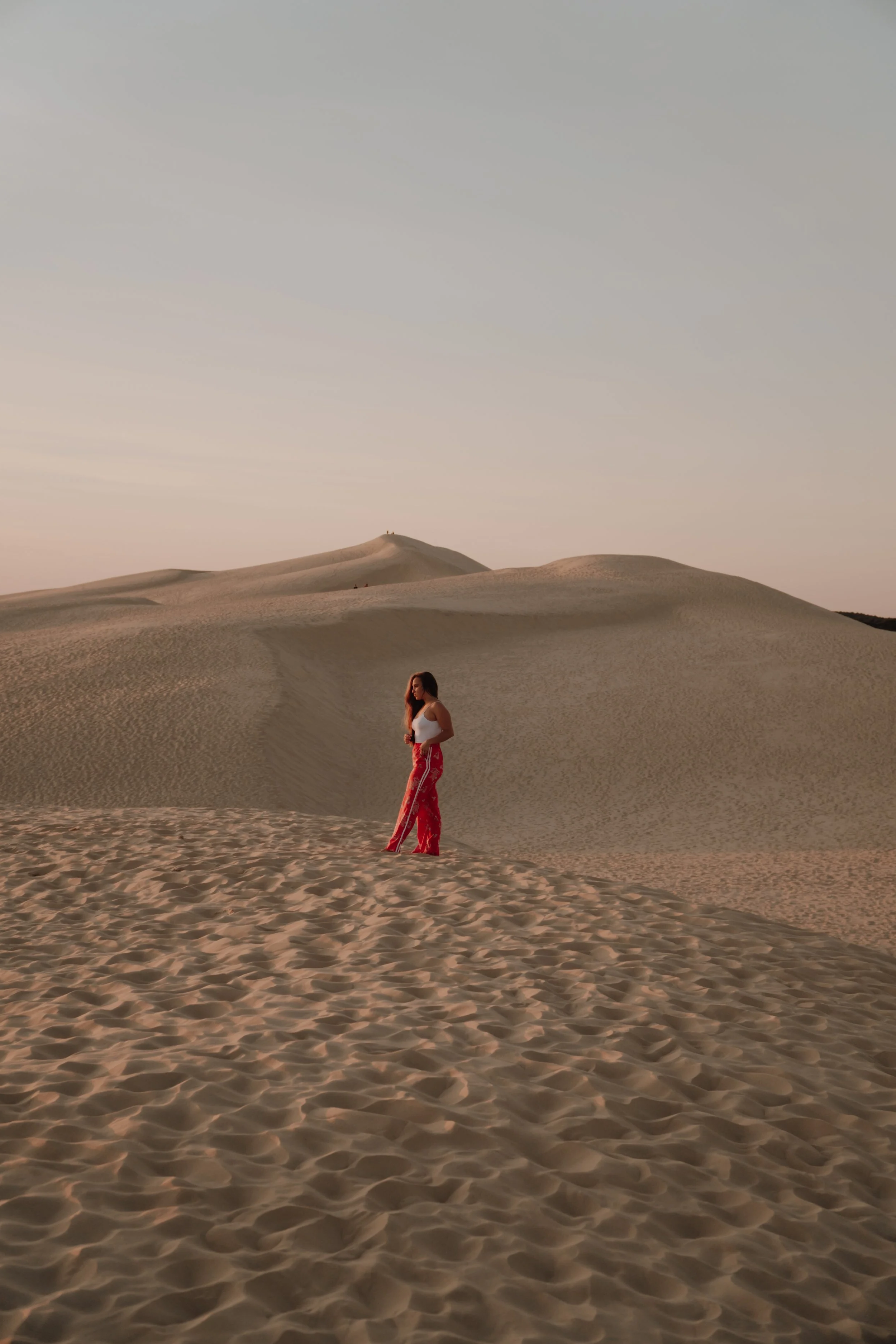 Une femme portant une robe rouge et un haut blanc se tient sur des dunes de sable dans un désert au coucher du soleil.