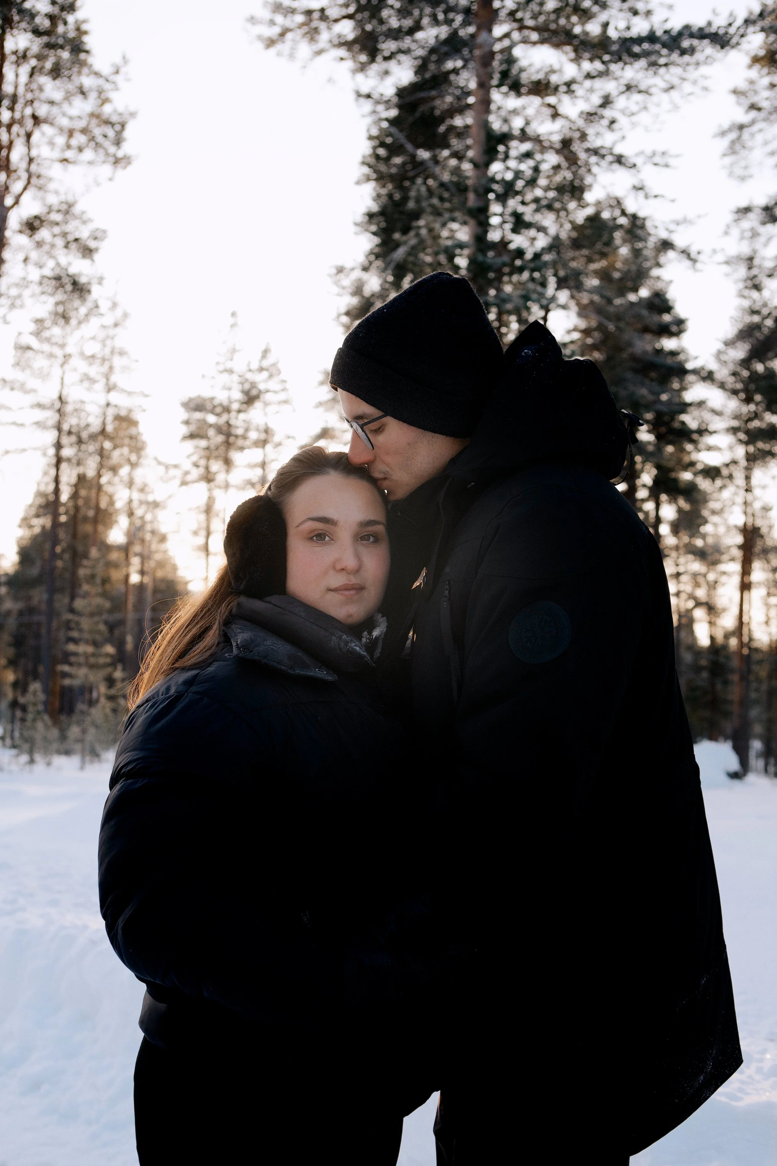 Un couple dans une forêt enneigée, l'homme embrasse tendrement la femme sur le front pendant que le soleil se couche à l'horizon