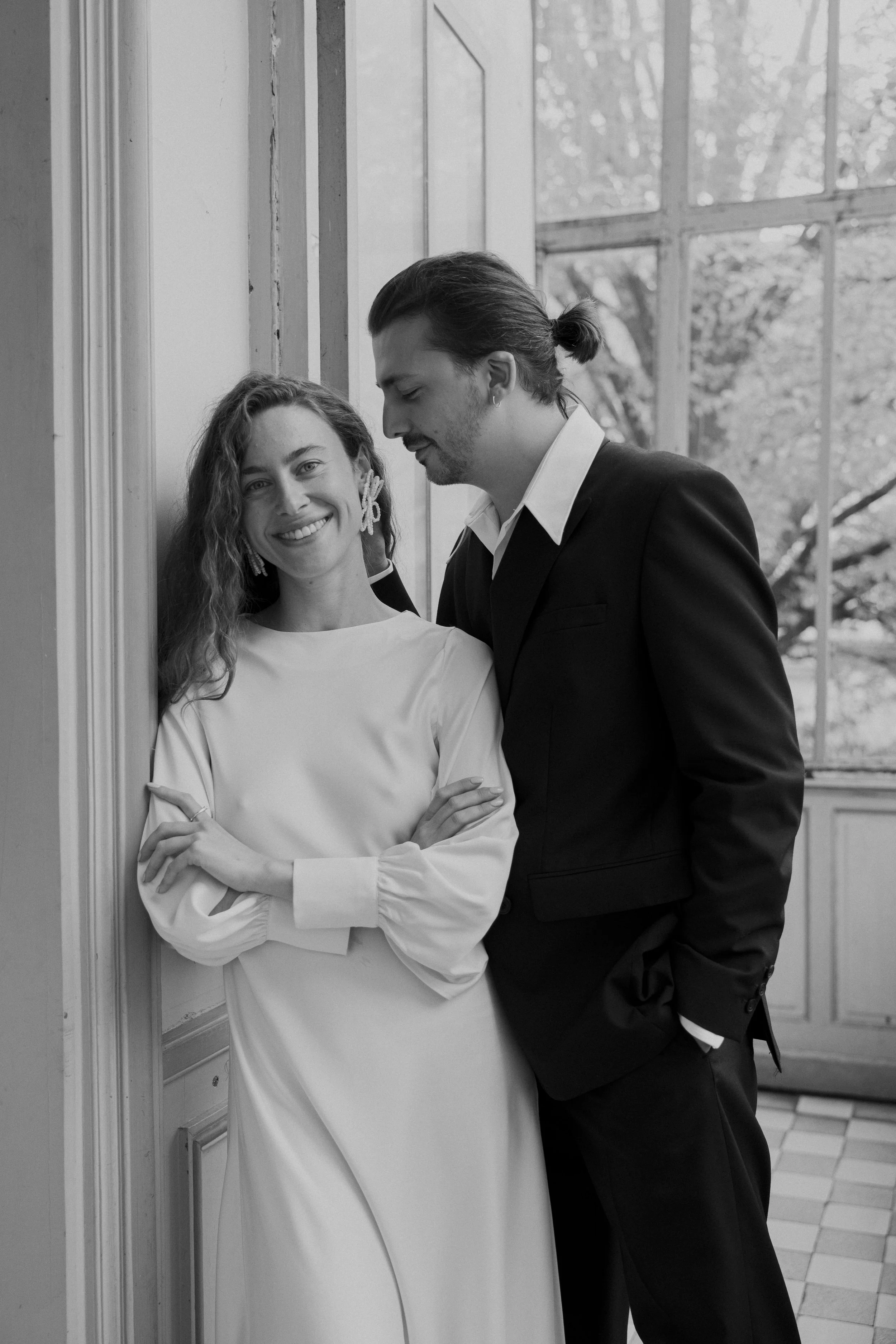 Un couple souriant en vêtements élégants, debout près d'un mur à l'intérieur, avec une grande fenêtre en arrière-plan montrant des arbres.
