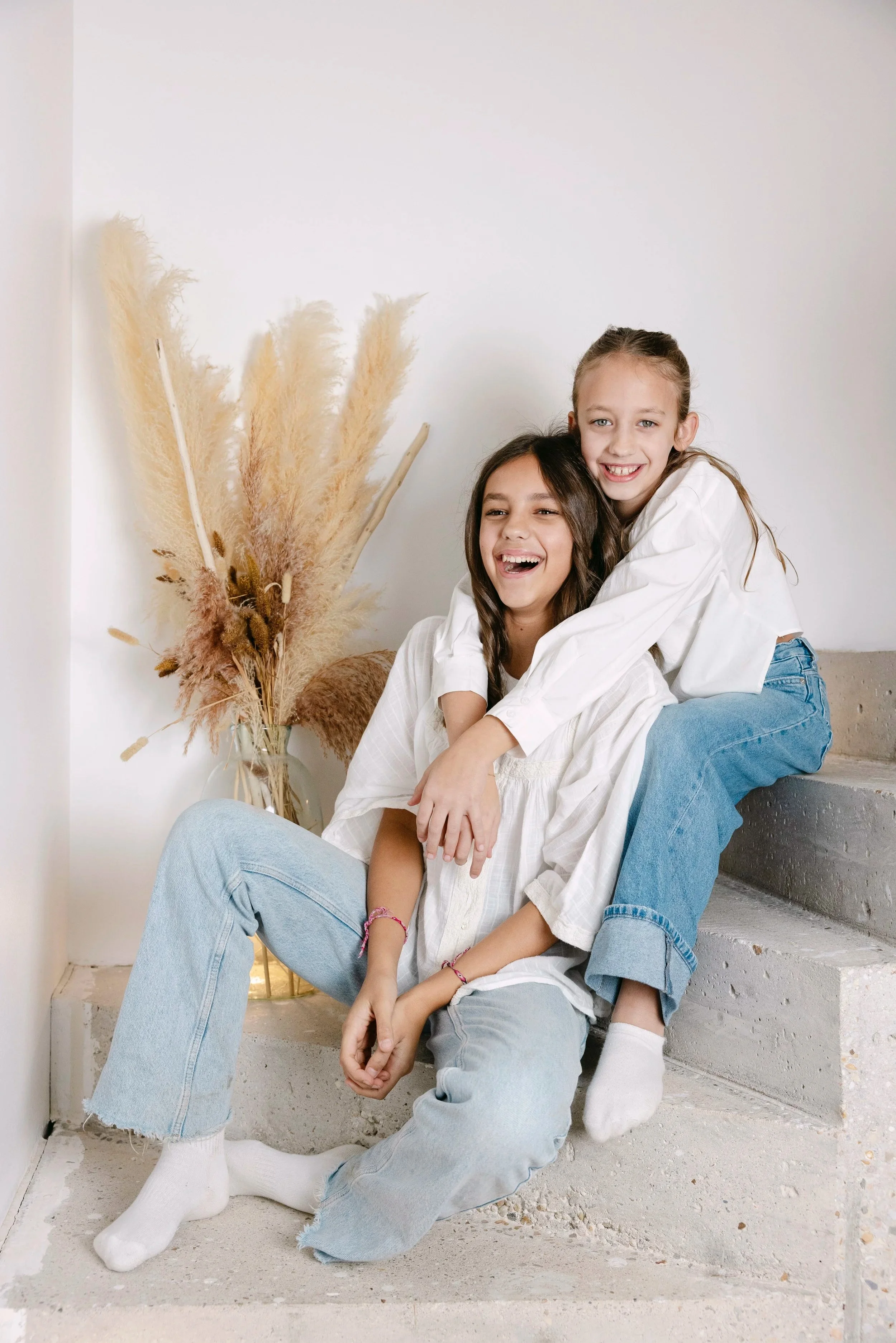 Deux jeunes filles souriantes et amusantes s'embrassent et jouent ensemble sur des escaliers en béton, dans une pièce minimaliste avec un vase de plantes séchées en arrière-plan.