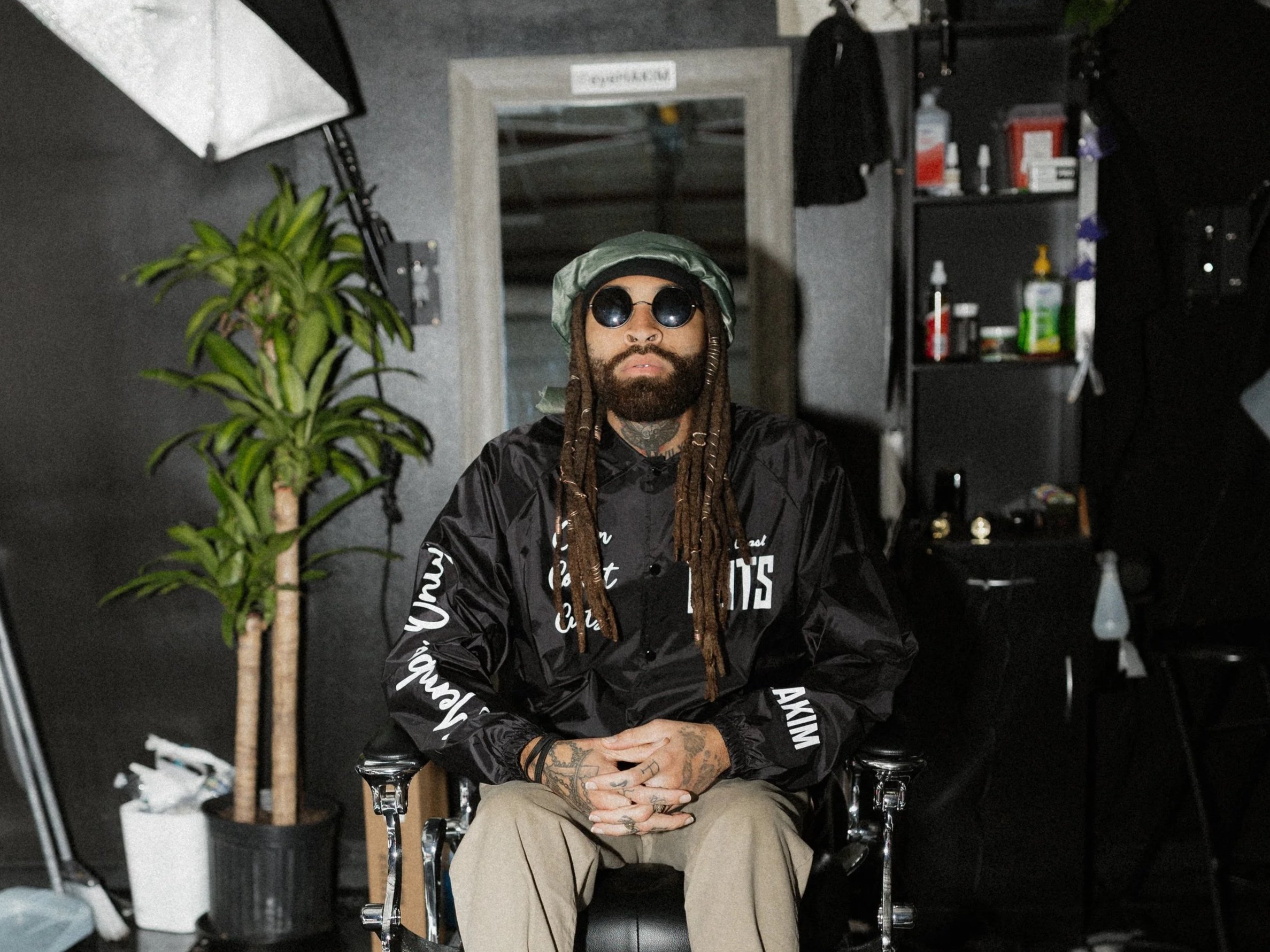 A man with long dreadlocks, beard, and sunglasses sitting in a tattoo chair with his hands clasped. He is wearing a black windbreaker jacket, beige pants, and a green bucket hat. The background shows shelves with various bottles and supplies, a mirror, a chalkboard, and a tall potted plant.