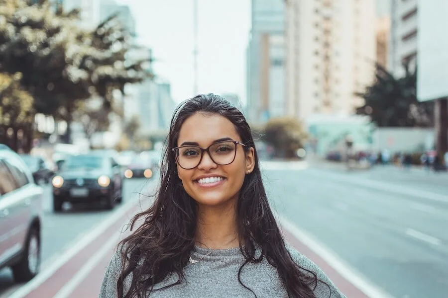Woman standing in the middle of a busy city street wearing glasses and smiling.