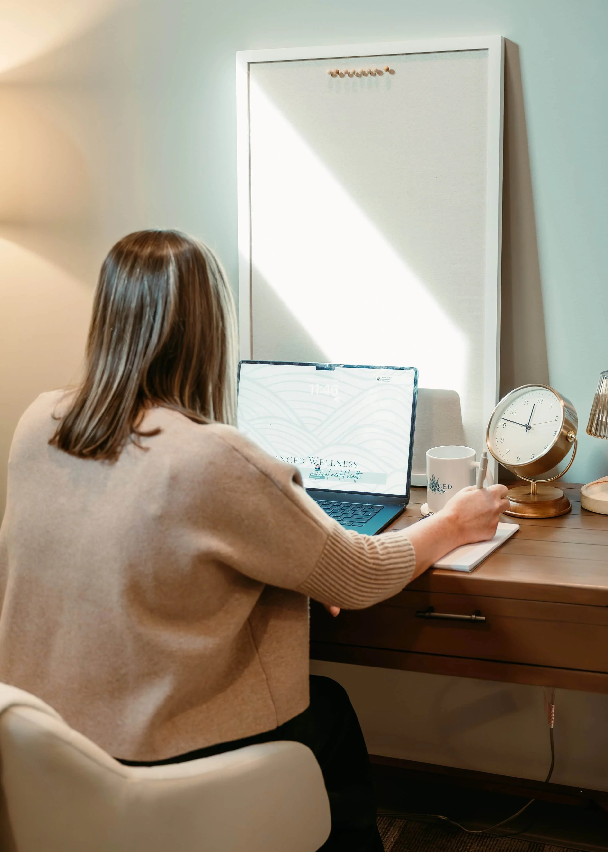 A woman sitting at her desk in front of a laptop writing in a notebook.