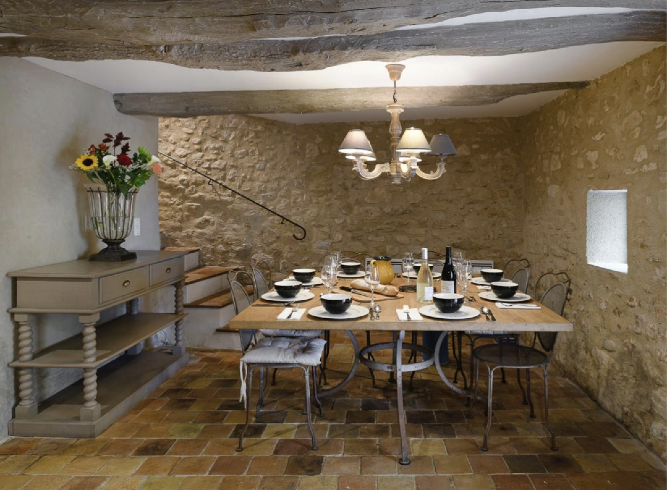 Dining room with a stone wall, wooden table set for dinner with black bowls, wine glasses, and wine bottles, surrounded by metal chairs, a chandelier with lampshades hangs from the ceiling, a small window, and a side table with a flower vase.