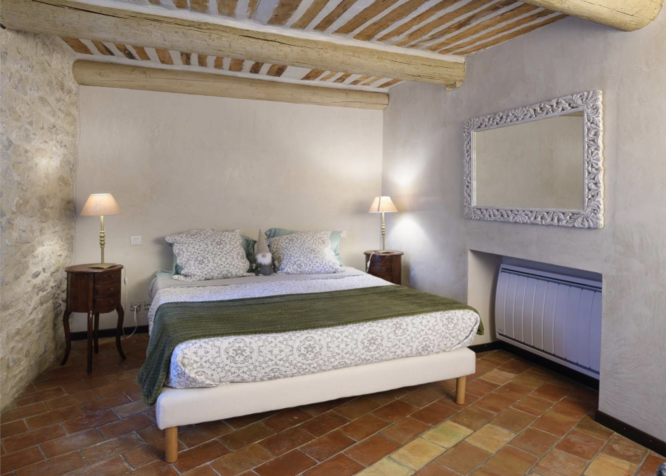 A cozy bedroom with a large bed, green and white bedding, and multiple pillows, flanked by two wooden nightstands with lamps, against a rustic wall with exposed brick and wooden beams on the ceiling.