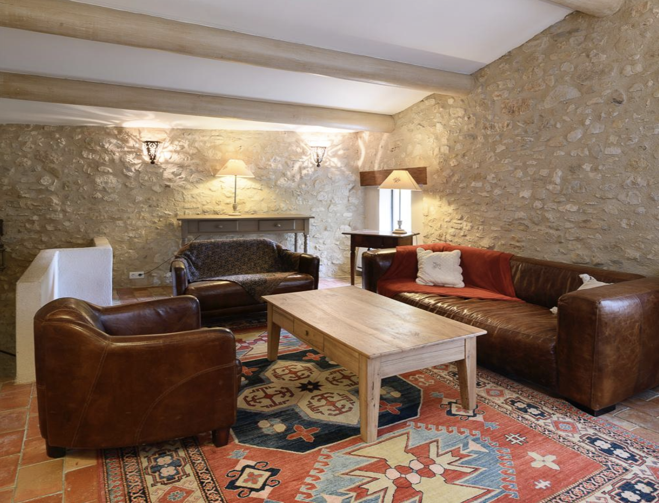 Cozy living room with a stone wall, wooden ceiling beams, leather and fabric sofas, a wooden coffee table, a patterned rug, and lamps providing warm lighting.