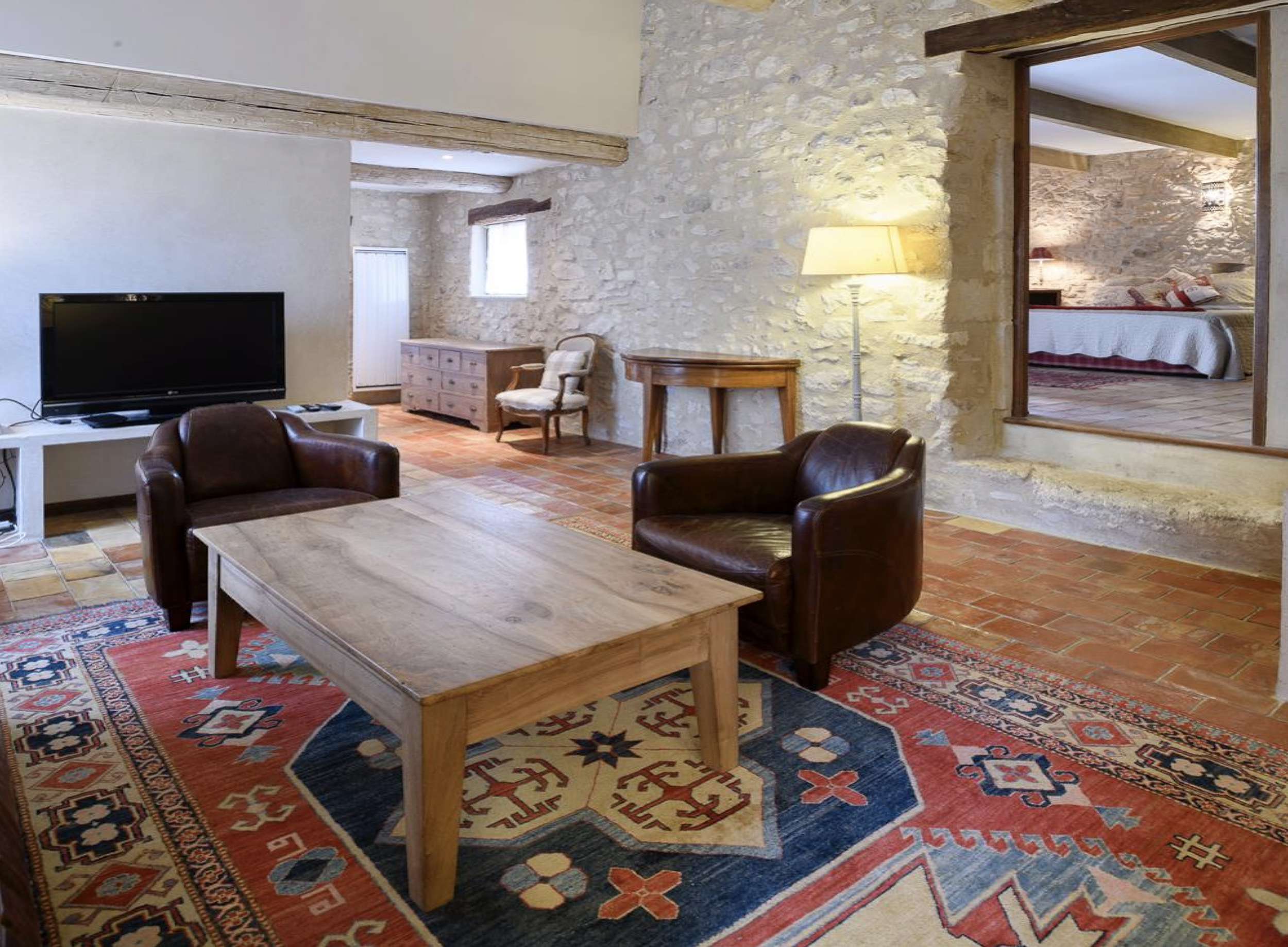 Living room with exposed stone walls, a large flat-screen TV, wooden coffee table, leather armchairs, patterned rug, and a view into a bedroom with a bed, pillows, and a nightstand.
