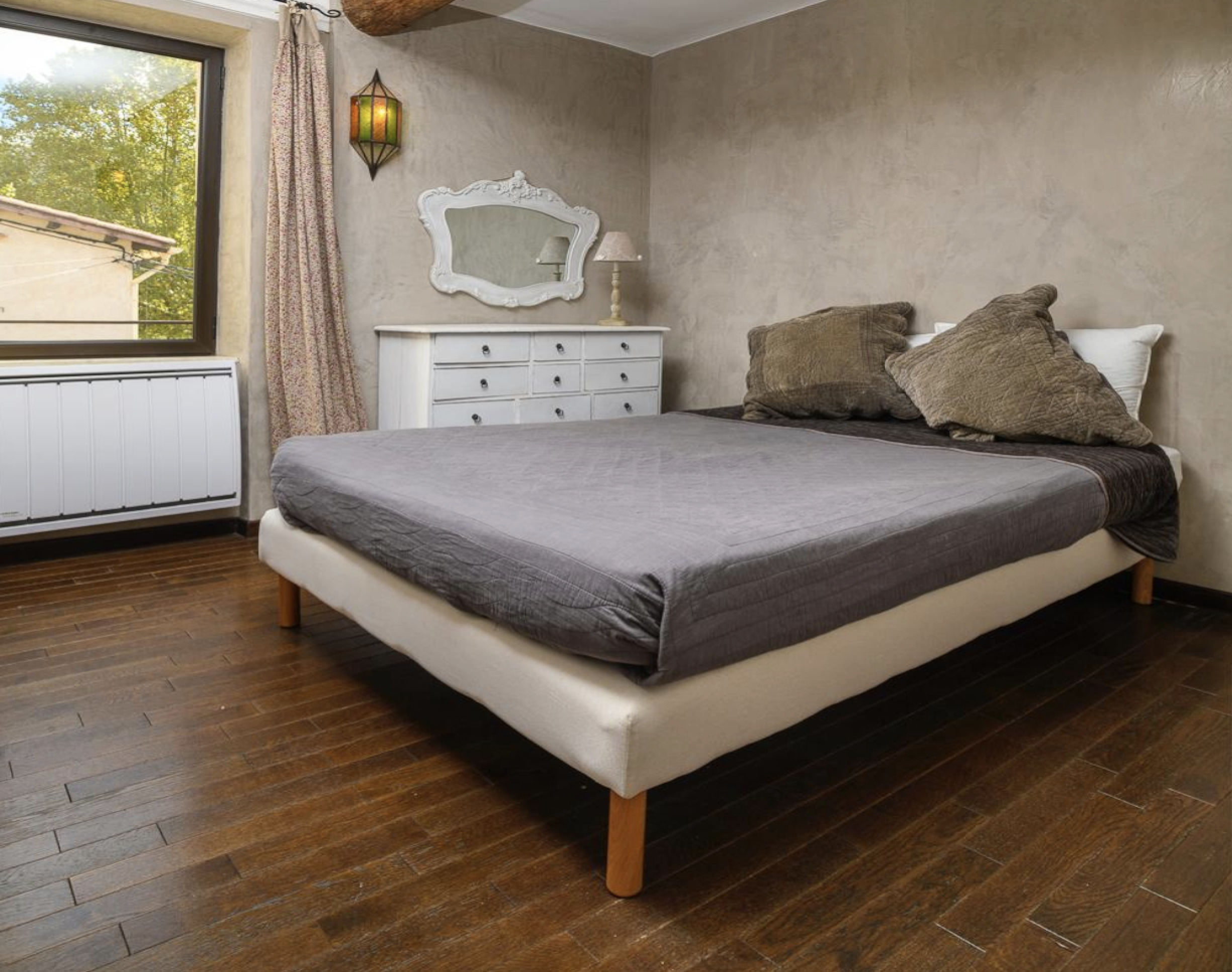 A bedroom with a bed that has brown pillows, a white dresser with a mirror, a lamp, a window with patterned curtains, and wood flooring.