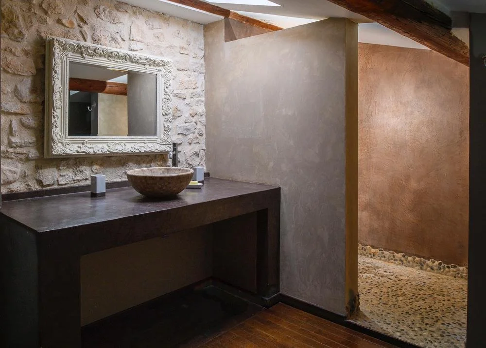Bathroom with stone and plaster walls, a black wooden countertop with a round stone vessel sink, a decorative framed mirror, and a pebble stone shower floor.