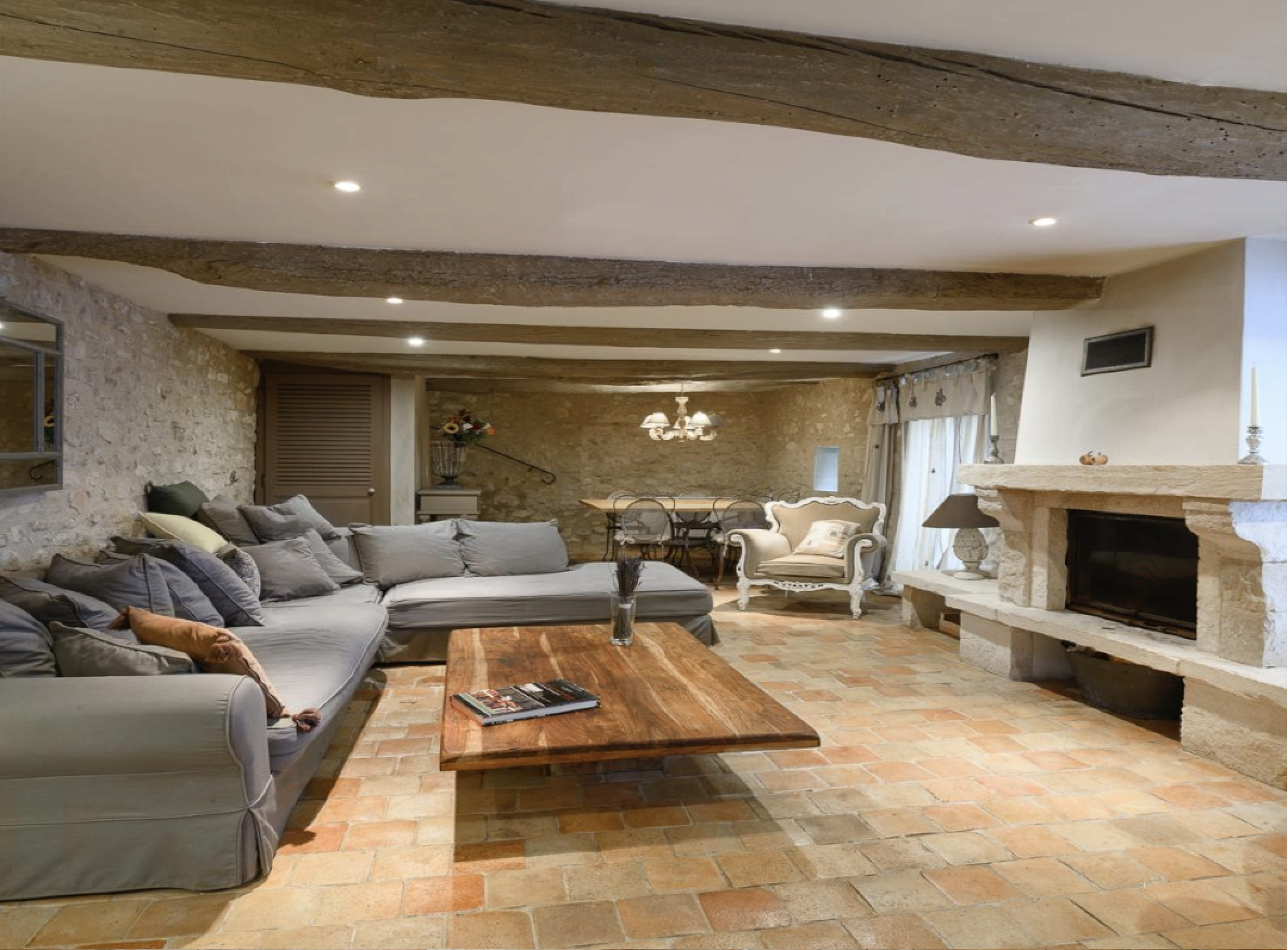 Air-conditioned living room with a gray sectional sofa, a wooden coffee table, an armchair, a fireplace, and a dining area with a table and chairs, decorated with flowers and curtains.