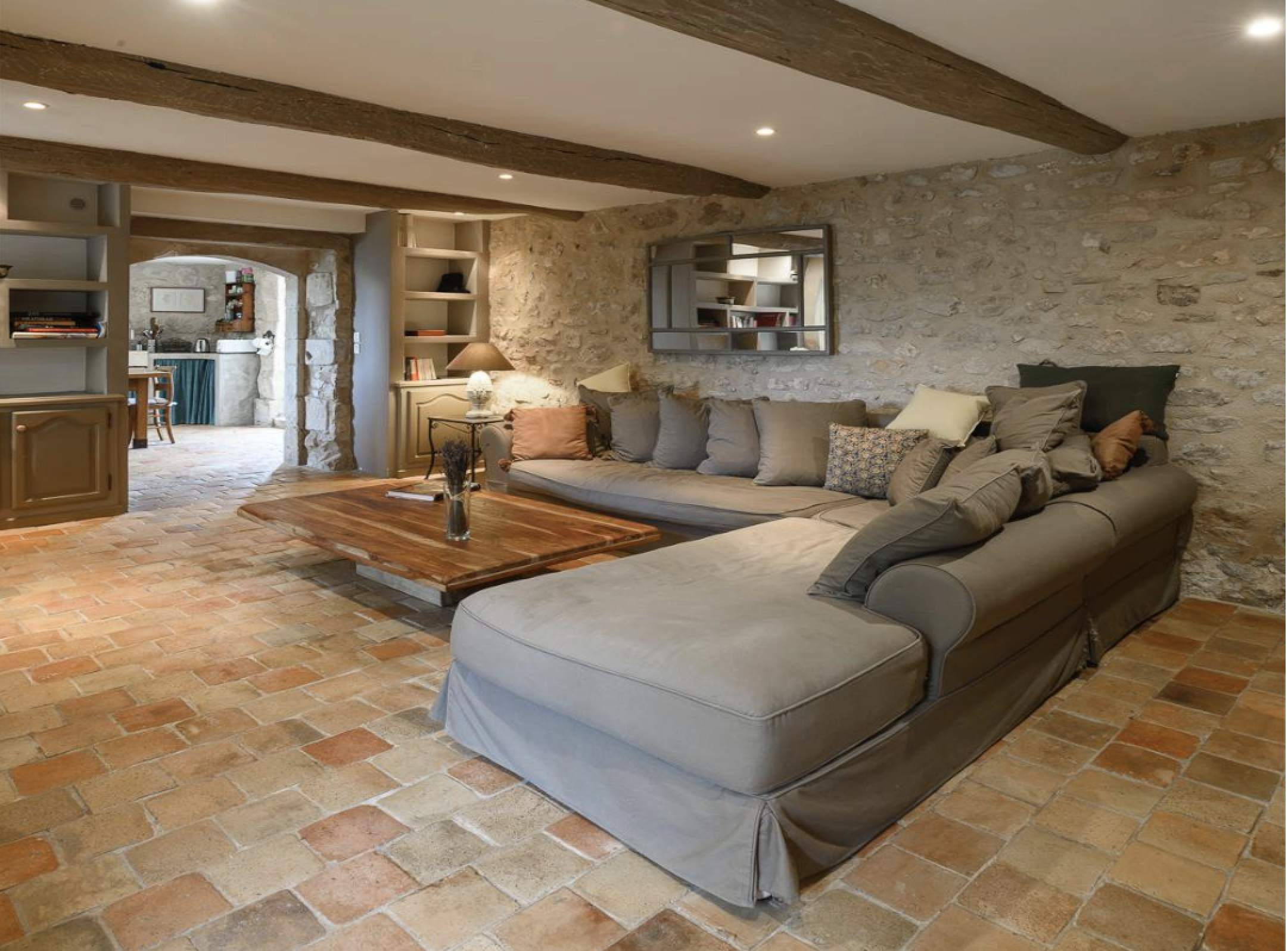 Living room with exposed stone wall, wooden ceiling beams, a beige sectional sofa with various pillows, a wooden coffee table, and brick flooring, with an open view into a kitchen area.