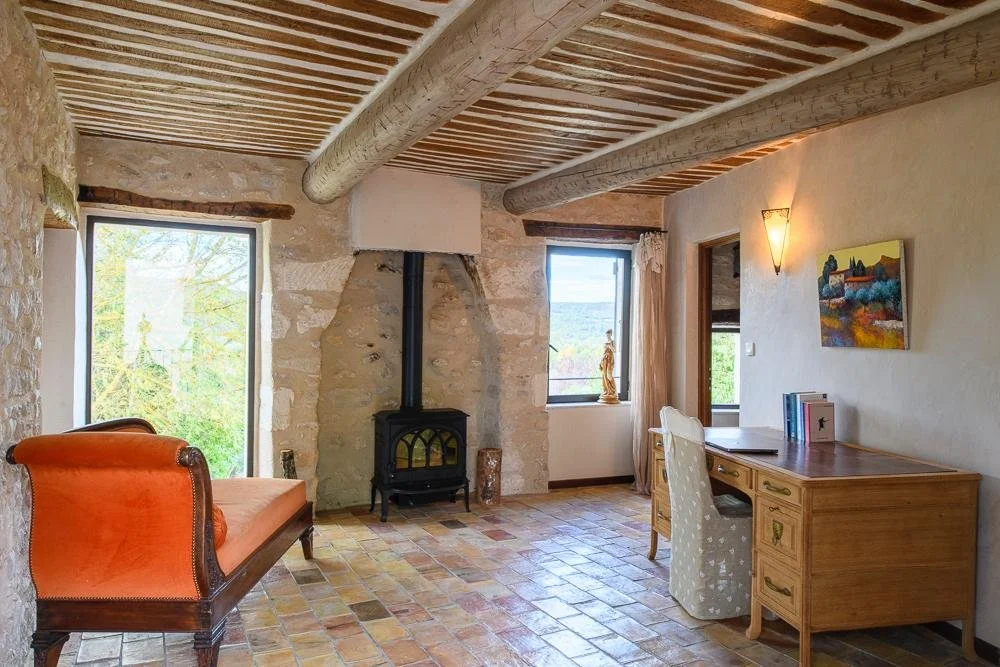 Room with stone walls, wooden ceiling beams, a small black wood stove, a sitting area with a vintage orange sofa, a desk with books, a window with a statue, another window, and a colorful landscape painting on the wall.