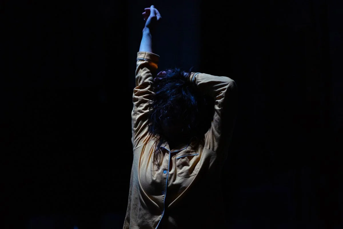 Person with dark hair in a beige shirt stretching with one arm overhead against a dark background.