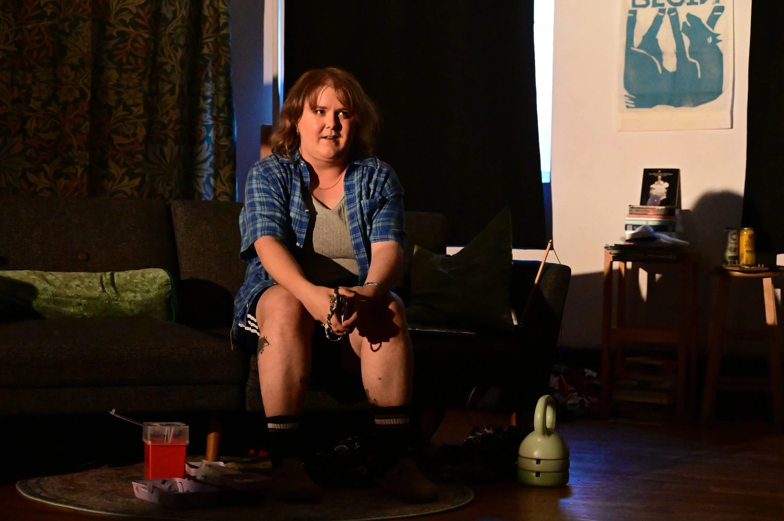 A woman with shoulder-length brown hair sitting on a dark brown couch in a dimly lit room. She is wearing a blue plaid shirt over a gray top and shorts, with her legs crossed. There is clutter on the floor in front of her, including a small clear container, some papers, and snacks. To her right, there is a small table with books, a framed photo, and other items. Behind her, a patterned curtain is visible on the left side and a poster or art piece on the wall to her right. There is also a green fan and some other small household items in the room.