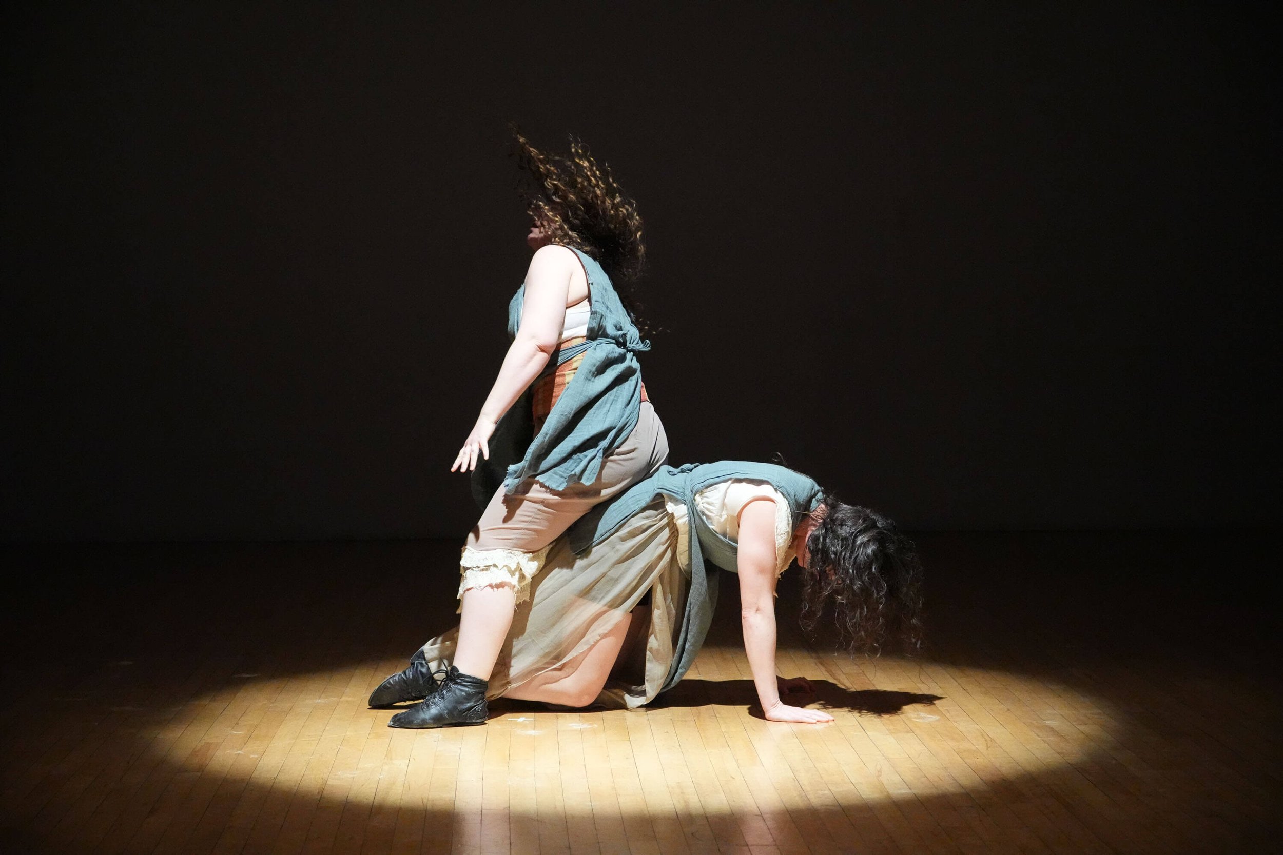 Two performers in costumes performing a dance or theatrical act on a stage with a dark background. One is on all fours, the other is kneeling and leaning forward with curly hair uplit.