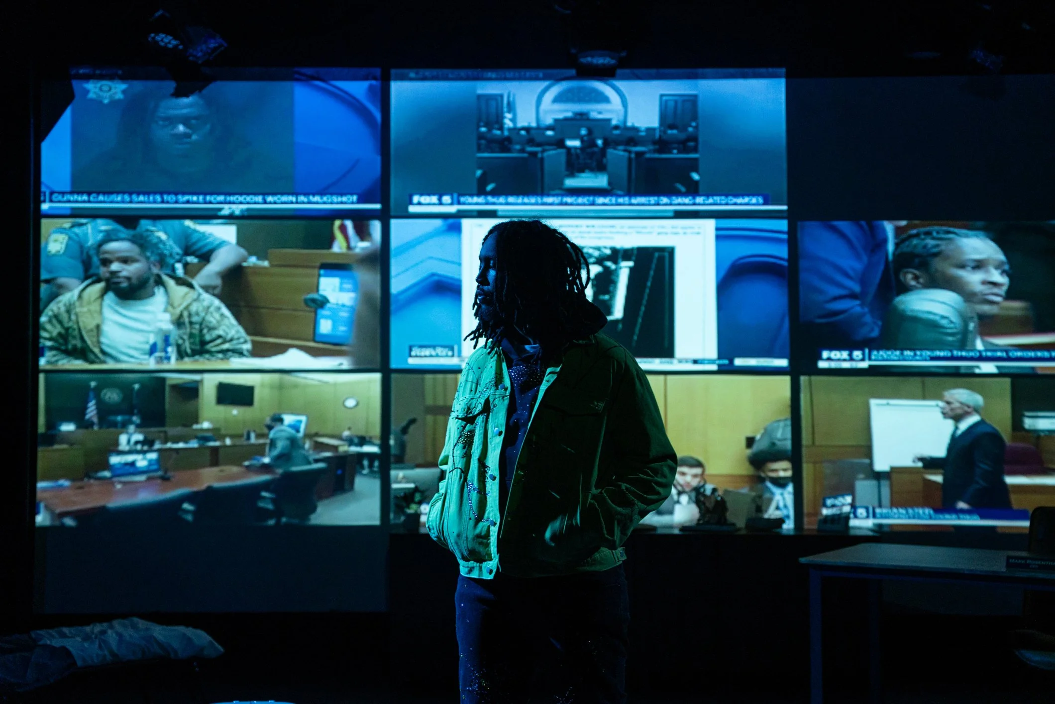 A person with long dreadlocks wearing a green jacket walking through a room with multiple large screens displaying news footage and images, including a courtroom scene and news anchors.