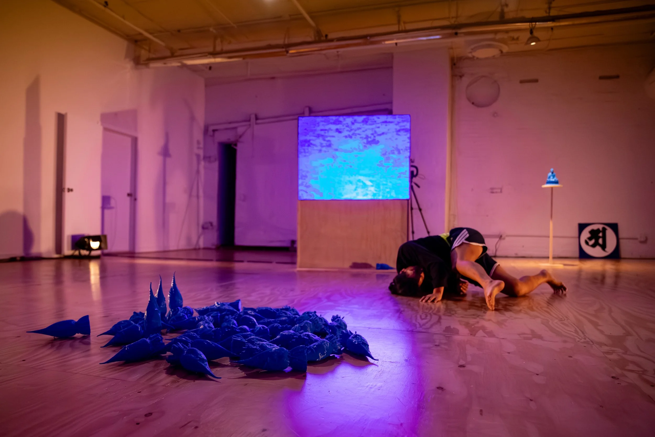 A dance performance with two performers on stage, one of whom is lying on top of the other, while many blue objects are scattered on the floor in front. There is a large digital screen displaying abstract visuals behind them, and soft theater lighting illuminates the scene.