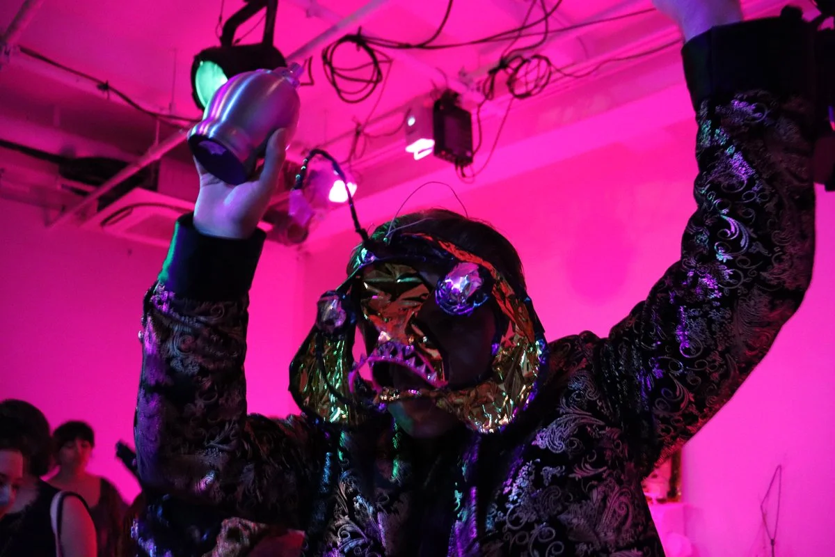 Person dressed in a reflective, patterned jacket wears a shiny mask and holds a metallic object in one hand, dancing under pink and purple lighting.