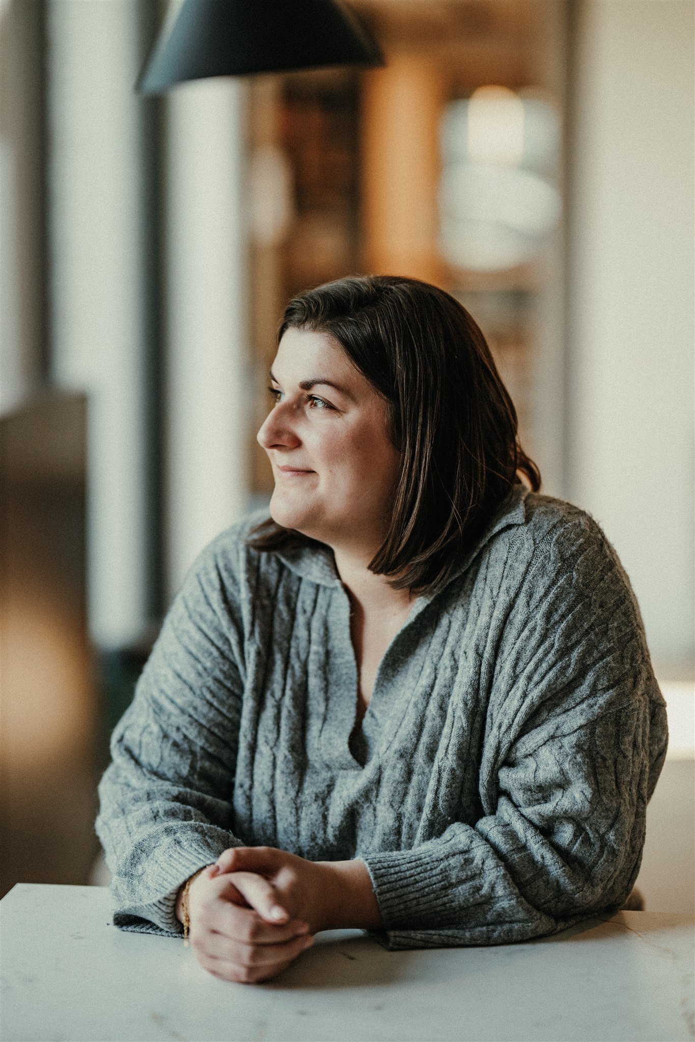 Een vrouw met schouderlang donker haar, zittend aan een tafel, kijkt naar links met een lichte glimlach in een huiselijke omgeving.