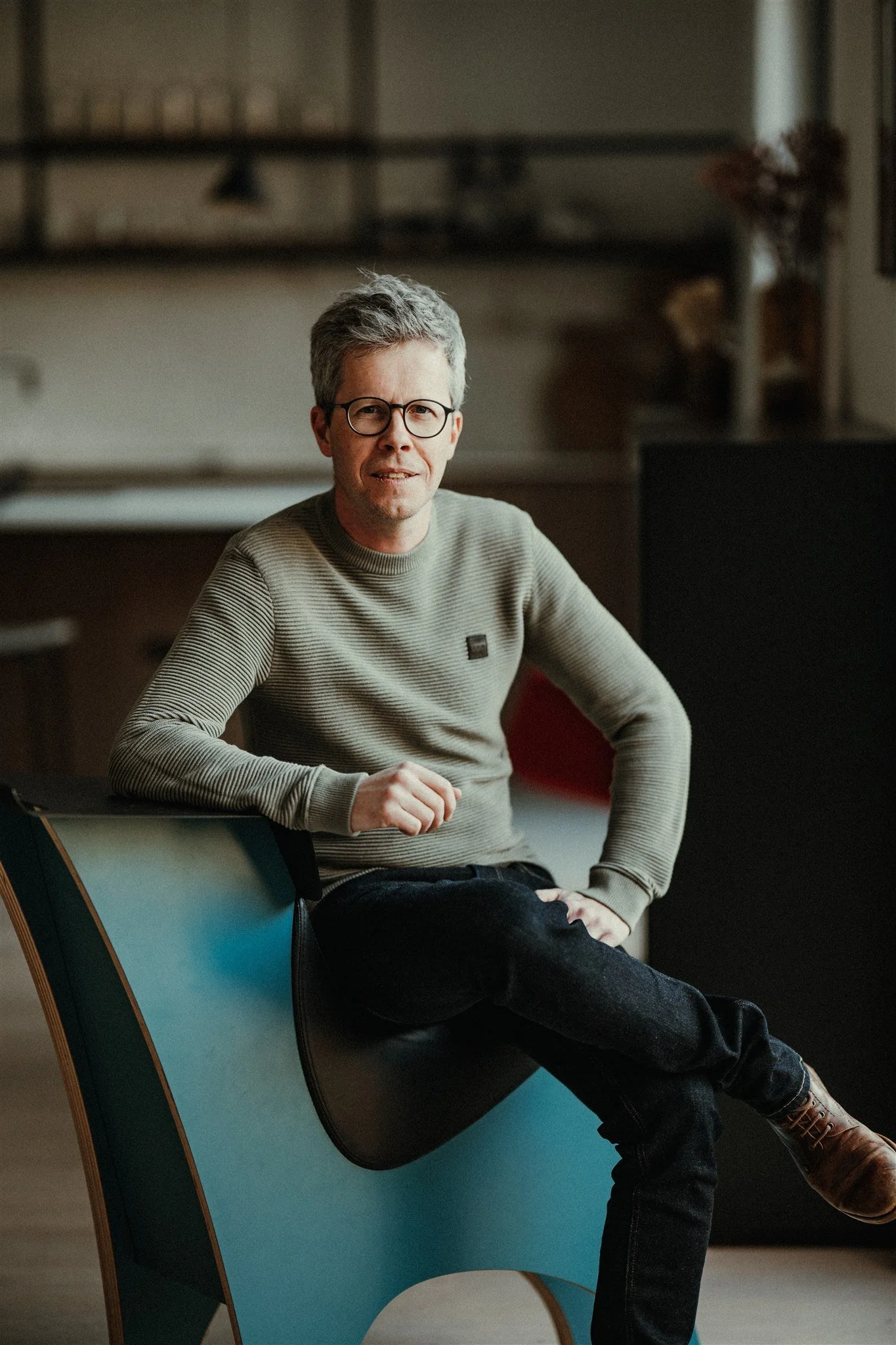 Man met grijze haar, bril en casual kleding zittend op een blauw ontwerpstuk in een interieur met boekenplank.