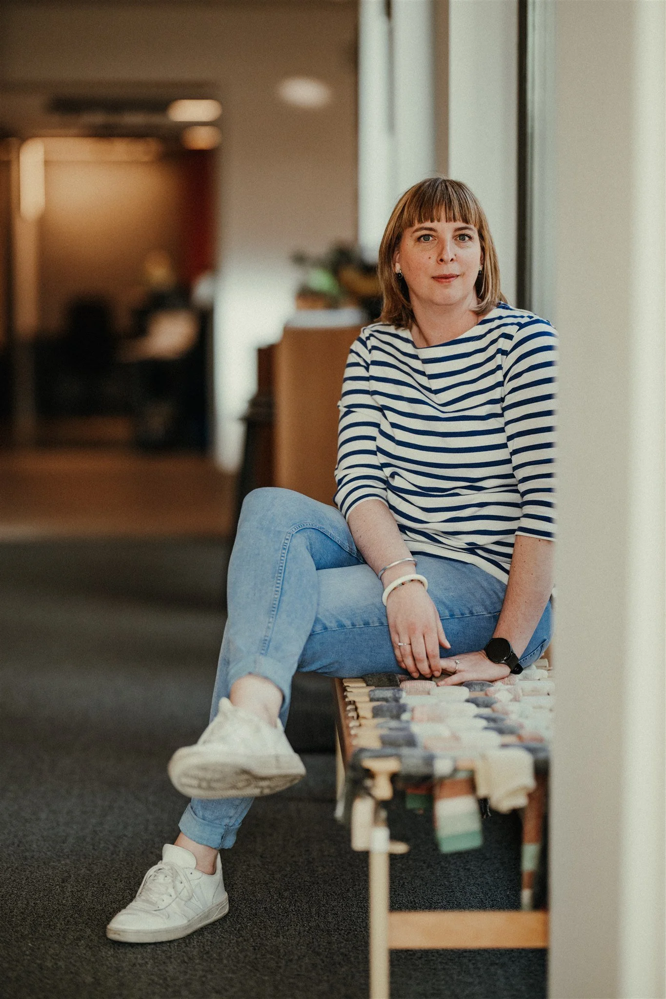 Vrouw zit op een bank, gekleed in een gestreept shirt en jeans, binnen bij een raam, met een achtergrond van een kamer.