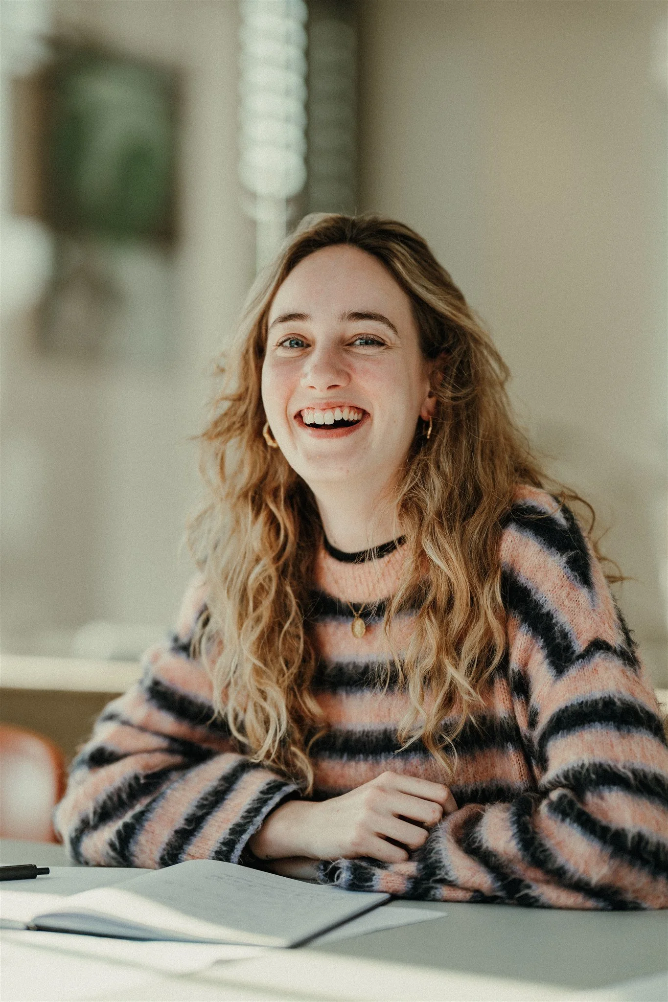 Een lachende vrouw met lang, krullend haar zit aan een tafel met een open notitieboekje en een pen.