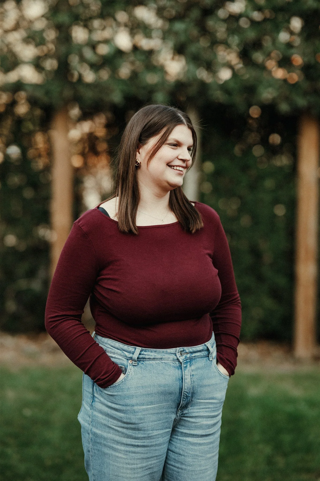 Een lachende vrouw met bruin haar, gekleed in een bordeauxrode trui en lichtblauwe jeans, staat buiten met een bosrijke achtergrond.