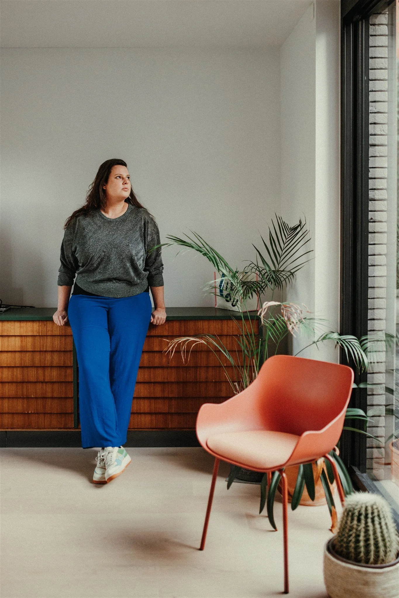 Vrouw leunend tegen houten dressoir in een modern interieur met planten en een oranje stoel.