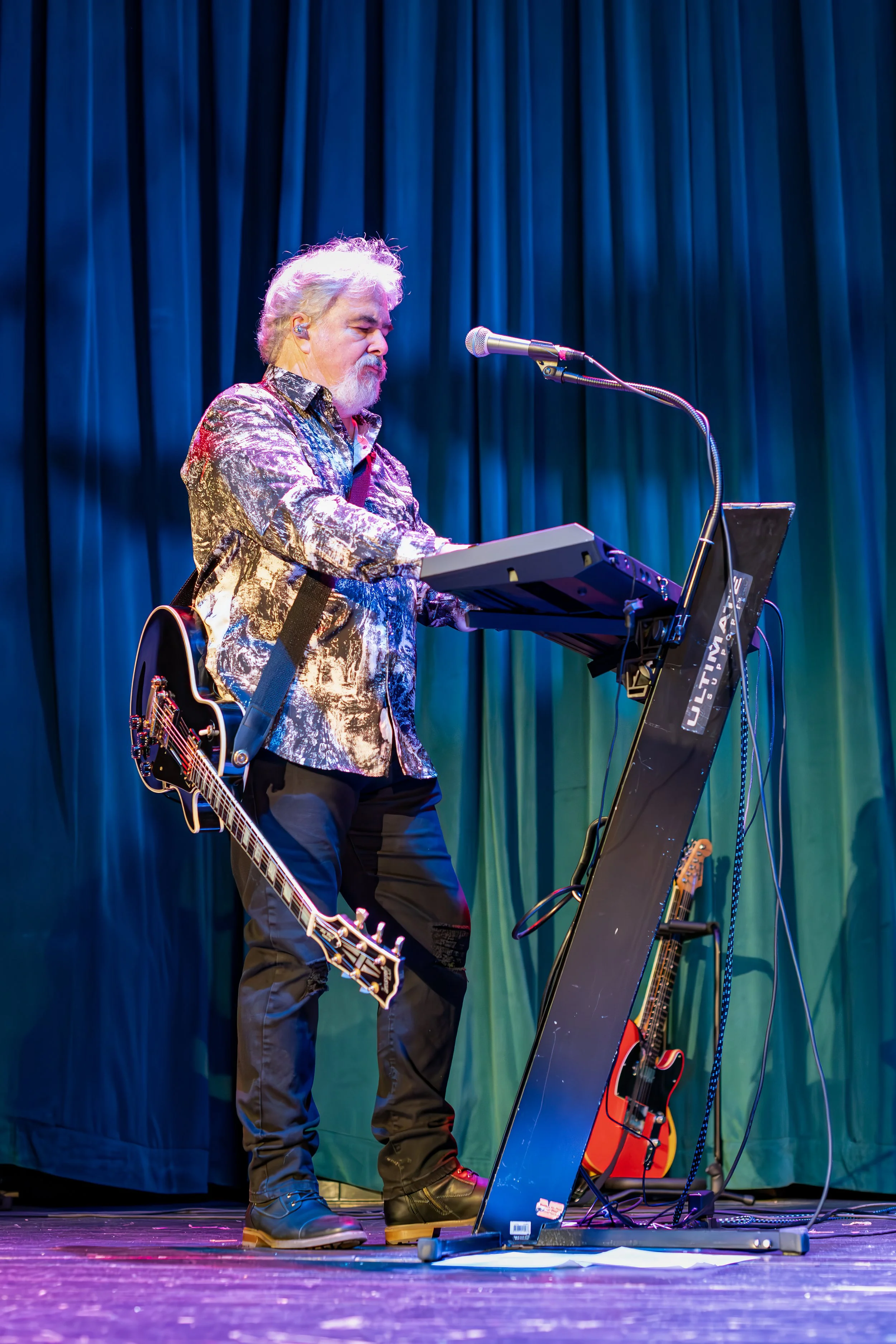 A musician performing on stage, playing keyboard with a guitar hanging from his shoulder, wearing a patterned shirt and black pants, with blue curtains in the background.