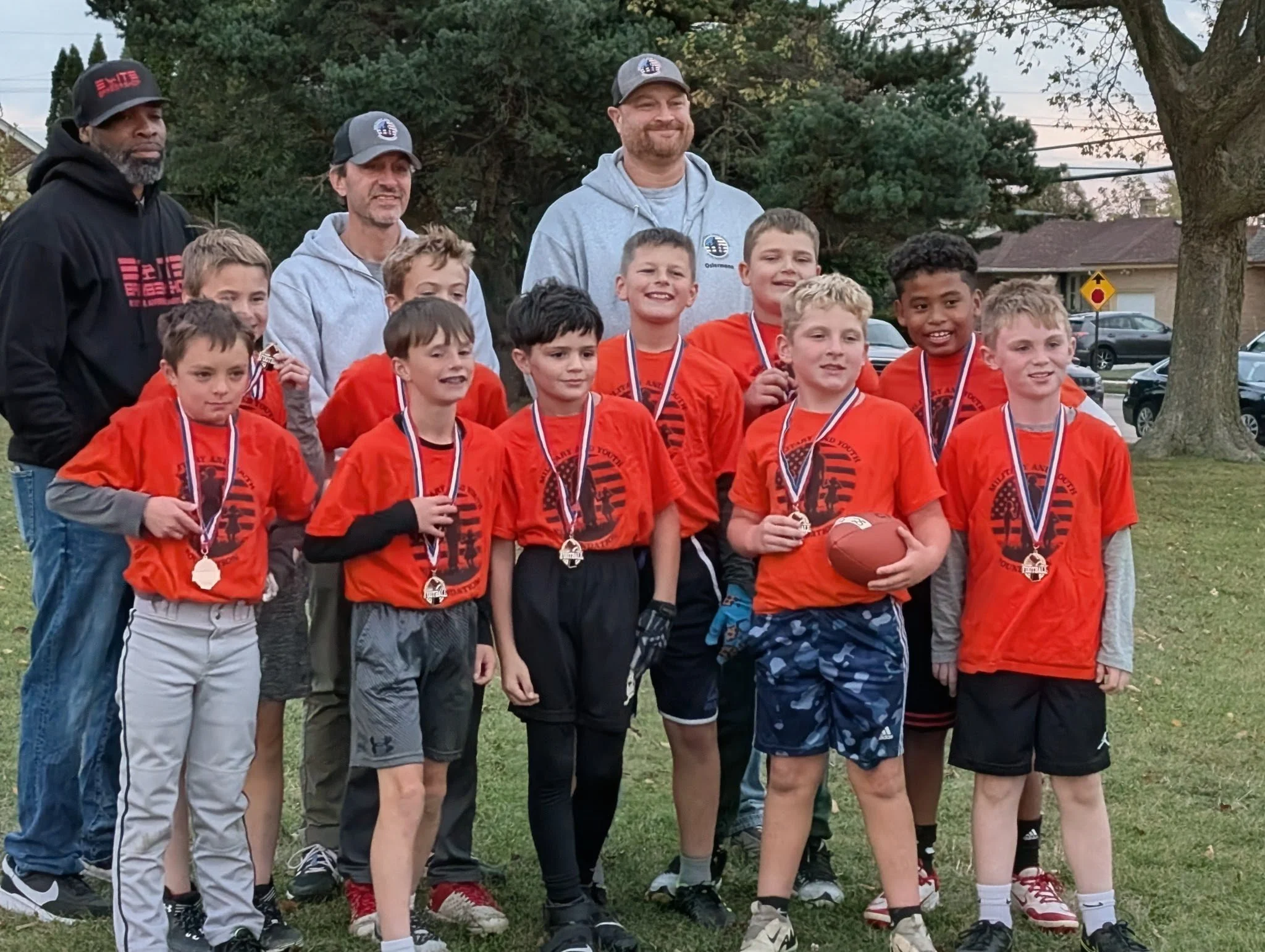 A group of boys in orange sports jerseys with medals around their necks, holding a football, standing outdoors in a park for a team photo, with adults and trees in the background.