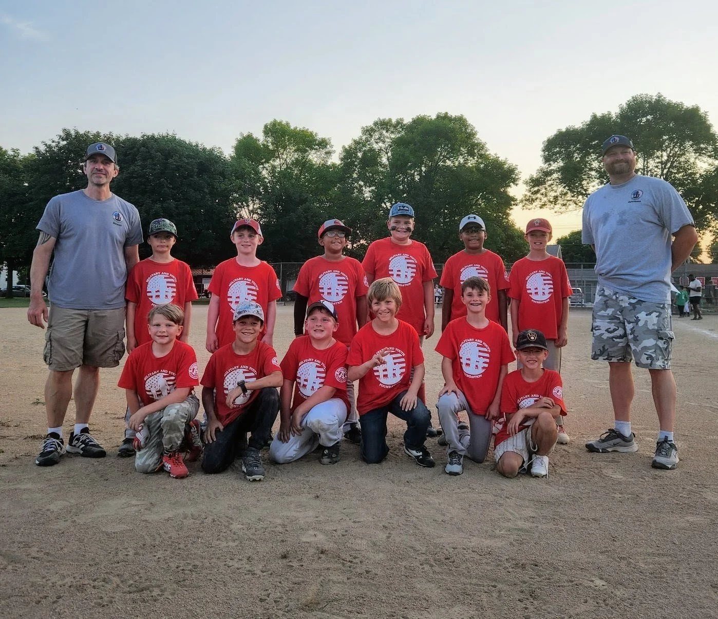 A youth baseball team with players and coaches posing on a baseball field at sunset.