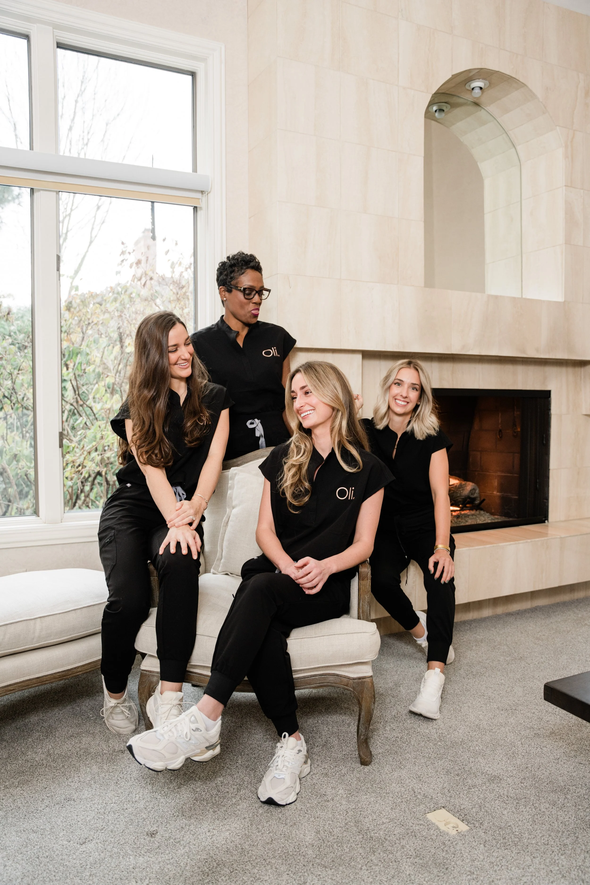 Four women sitting and standing together in a living room with a large fireplace, wearing black scrubs with the word 'Oli' on them, smiling and engaging with each other.