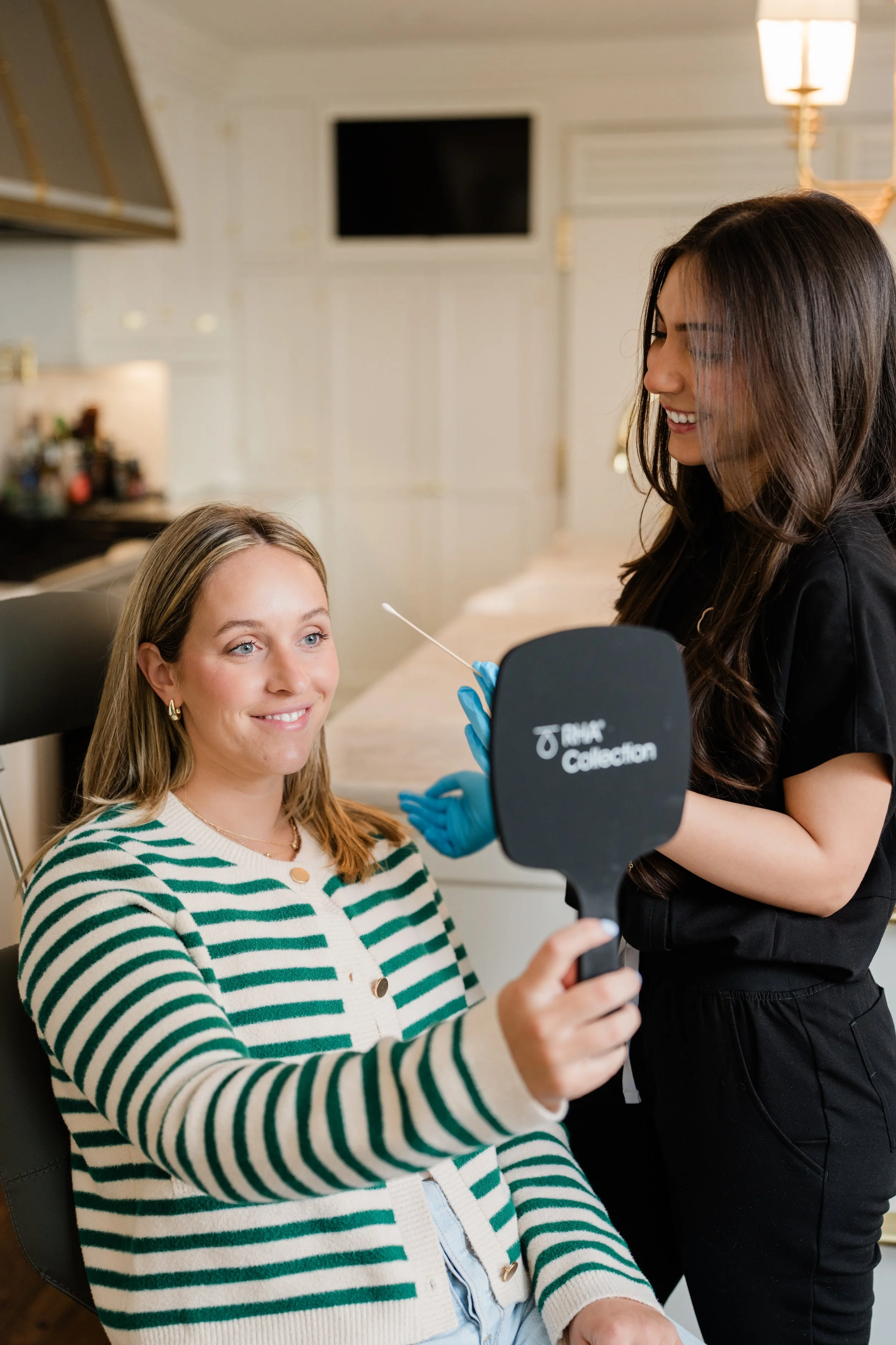 A woman with glasses and a striped sweater sitting in a dental chair, holding a mirror and smiling while a dental technician in black scrubs and gloves performs a dental procedure with a dental tool in a modern dental clinic.