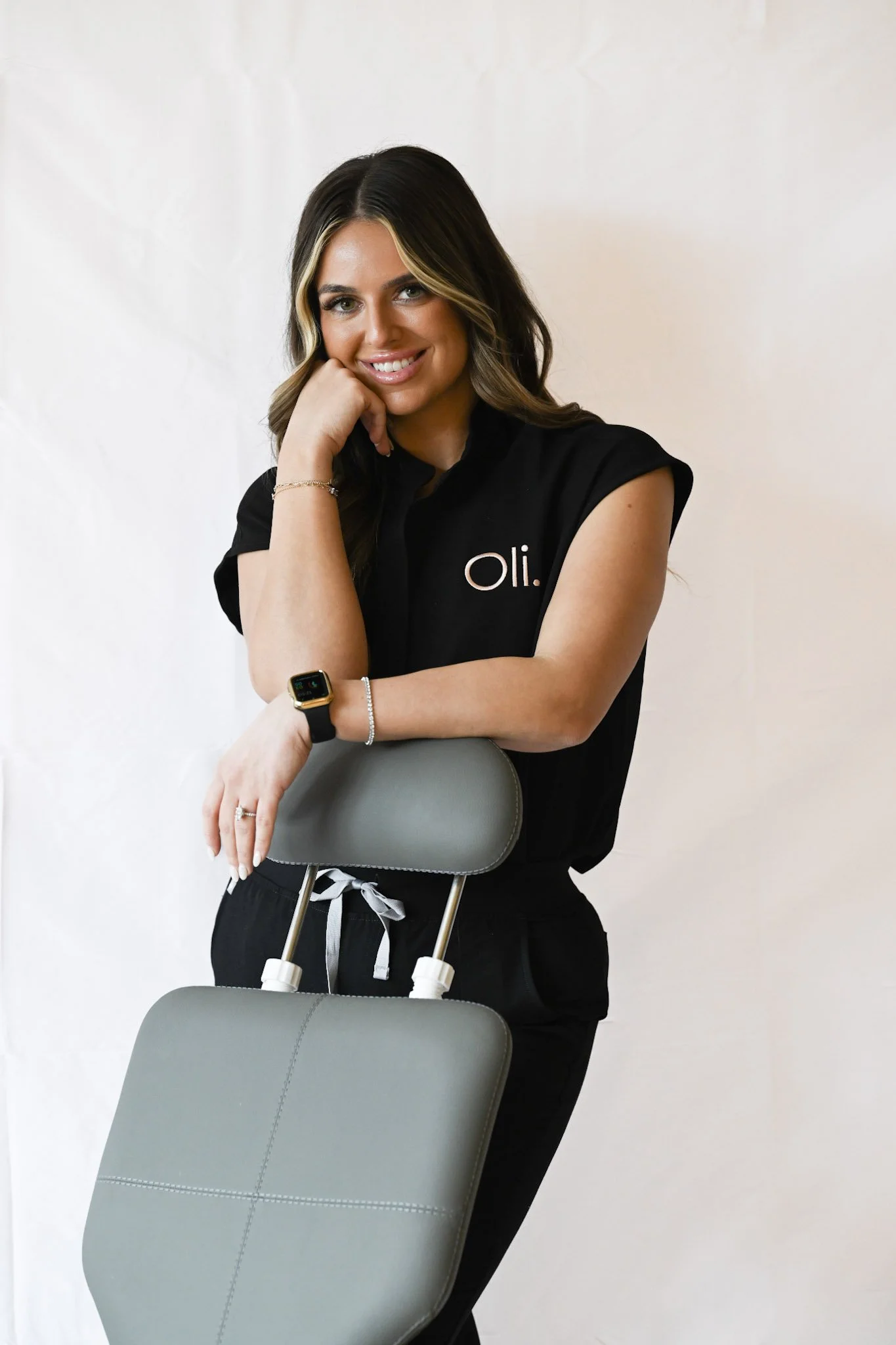 A smiling woman with wavy brown hair and light skin, wearing a black shirt with white text 'oli.' on the chest, and black pants. She is standing against a plain background, holding a gray massage or medical chair with her right hand, wearing a smartwatch and a bracelet on her left wrist, and resting her head on her left hand.