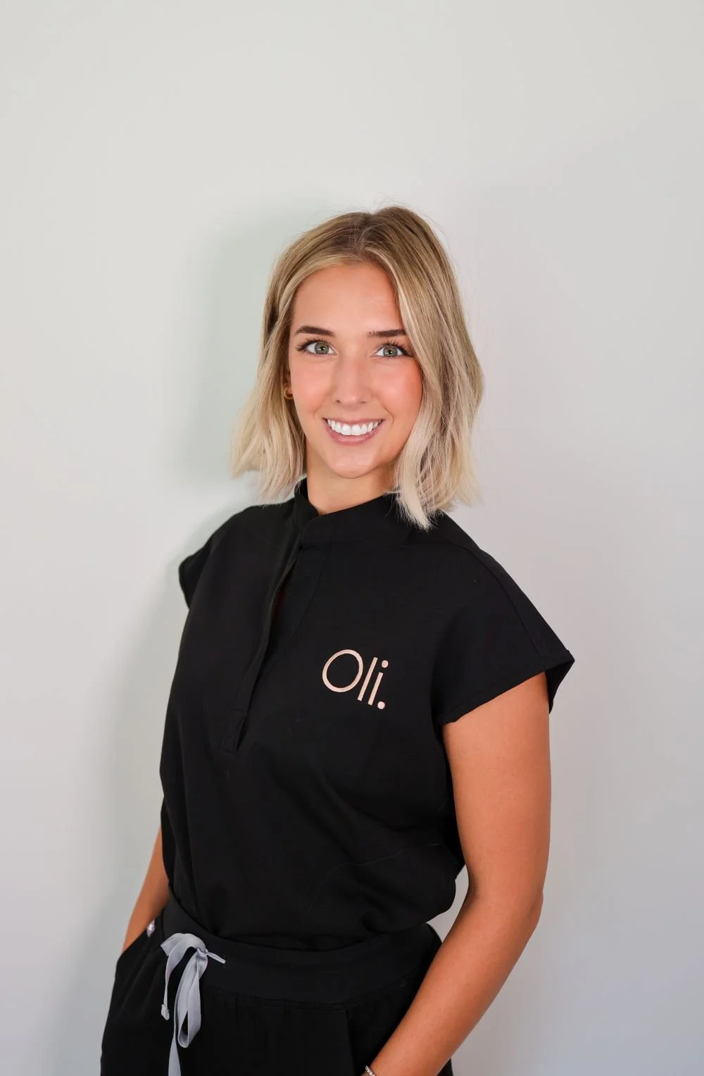 A young woman with shoulder-length blonde hair, wearing a black work uniform with 'oil:' written on it, smiling against a plain light-colored background.