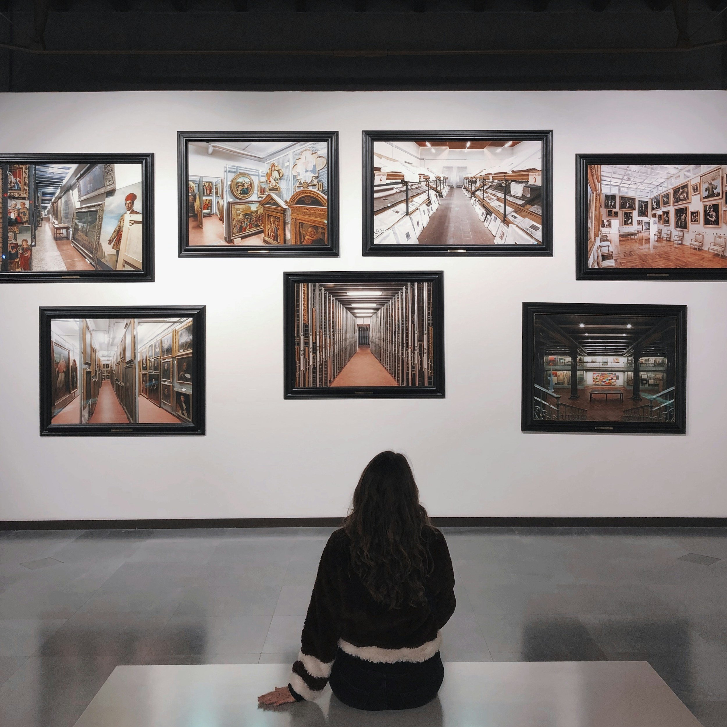 A person sitting on the floor in a gallery or museum, facing away from the camera, with long dark hair and wearing a black and white striped sweater. They are looking at framed artwork on a white wall.